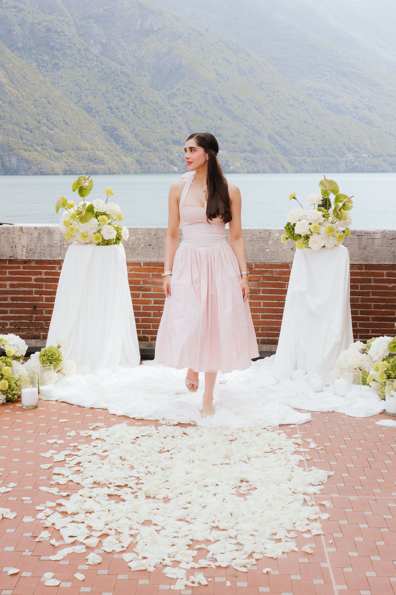 Bride in pink midi dress stands among white rose petals on terrace with floral arrangements and mountain lake backdrop.