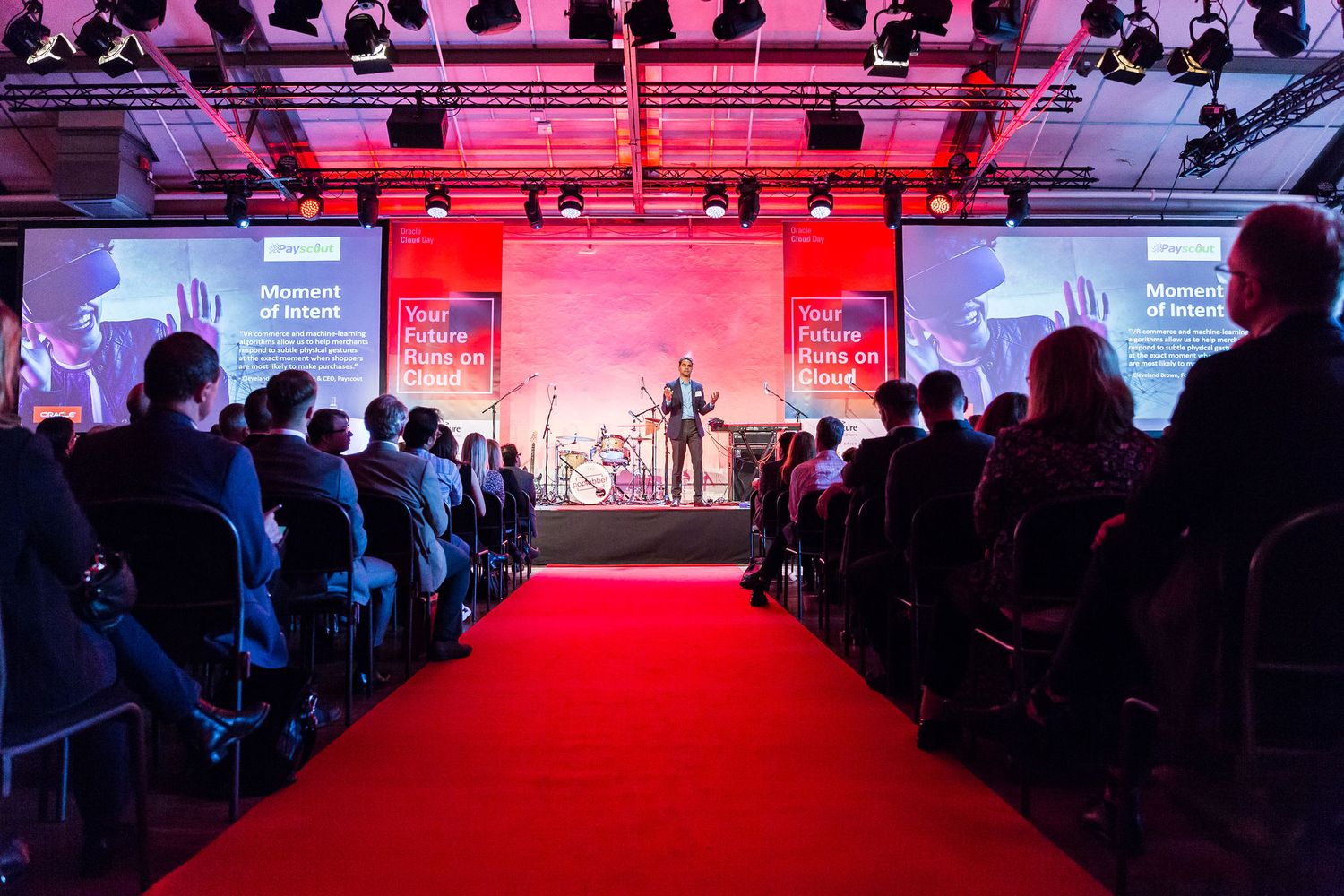 Speaker presenting on stage at a corporate event with red carpet aisle and seated audience under dramatic red lighting.