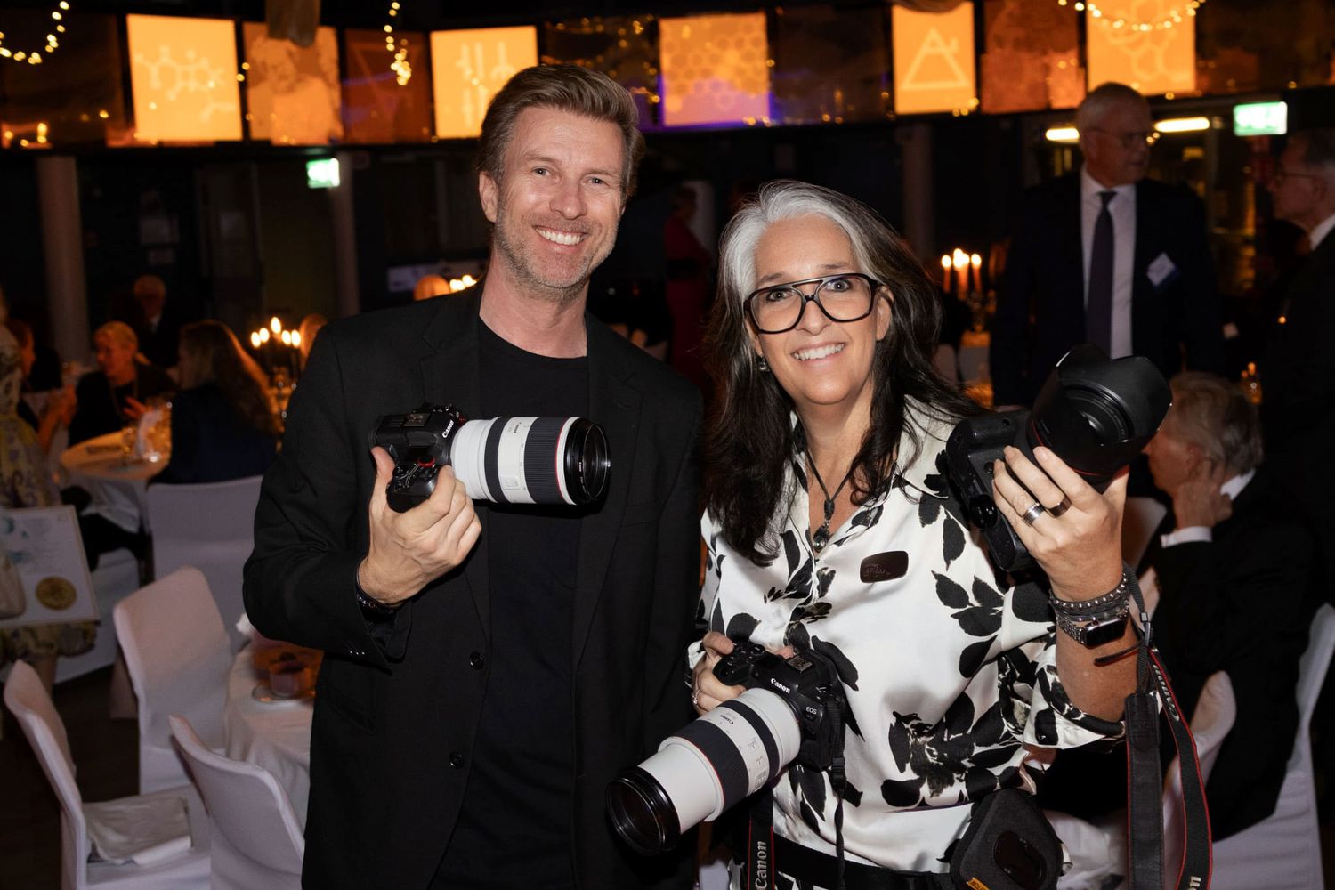 Two photographers smiling and holding professional cameras at an elegant indoor event venue.