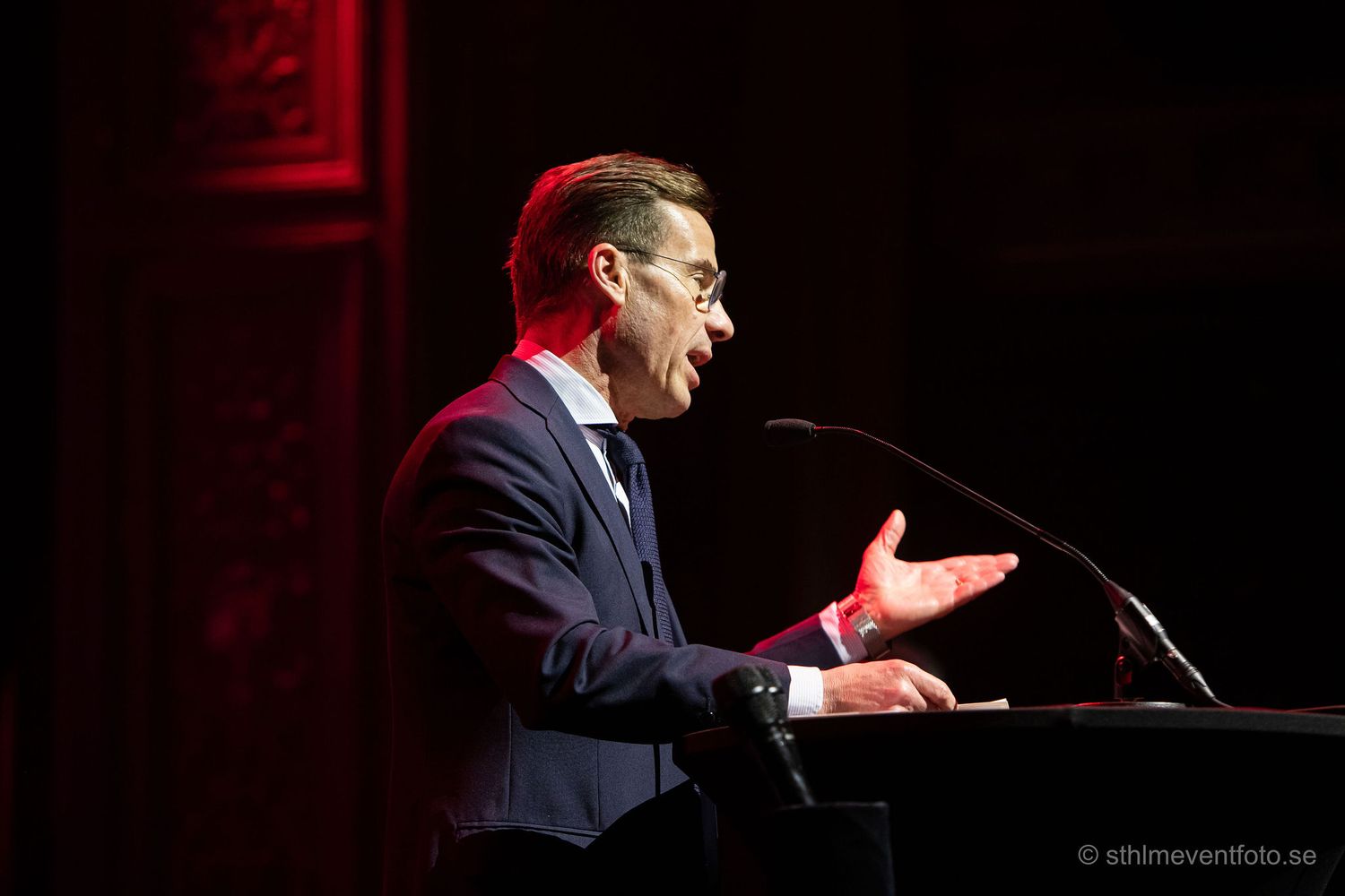 Man in navy suit speaking passionately at podium with microphone on stage under dramatic red lighting.