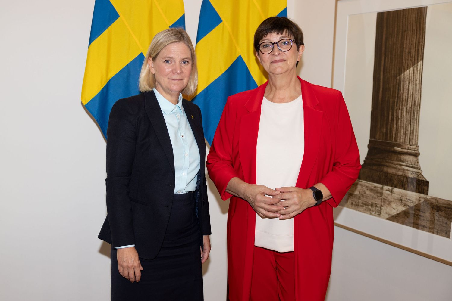 Two women pose together in front of Swedish flags, one in a black suit and one in a red blazer.