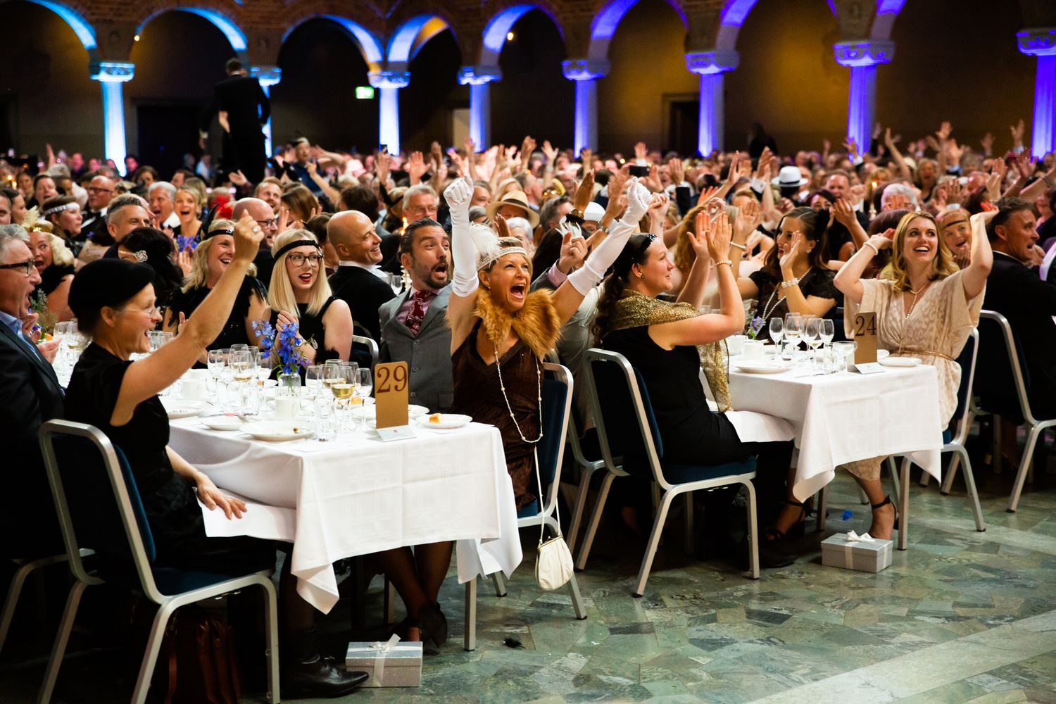 Large crowd of excited guests celebrating at a formal gala dinner event, raising hands and cheering at banquet tables.