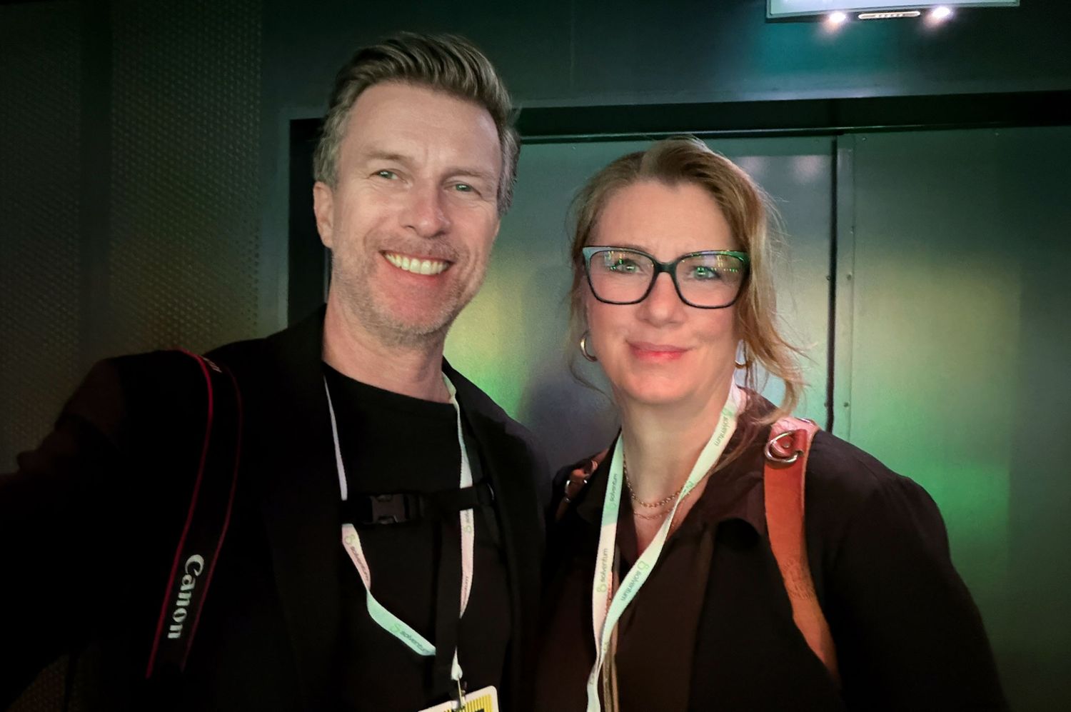 Two smiling professionals wearing lanyards pose together at an indoor event, man with Canon camera strap.