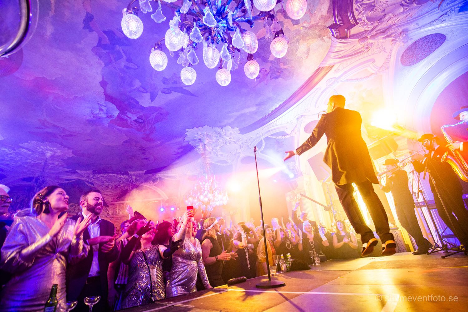 Performer on stage energizes crowd in ornate ballroom with blue and purple lighting and elegant chandeliers.