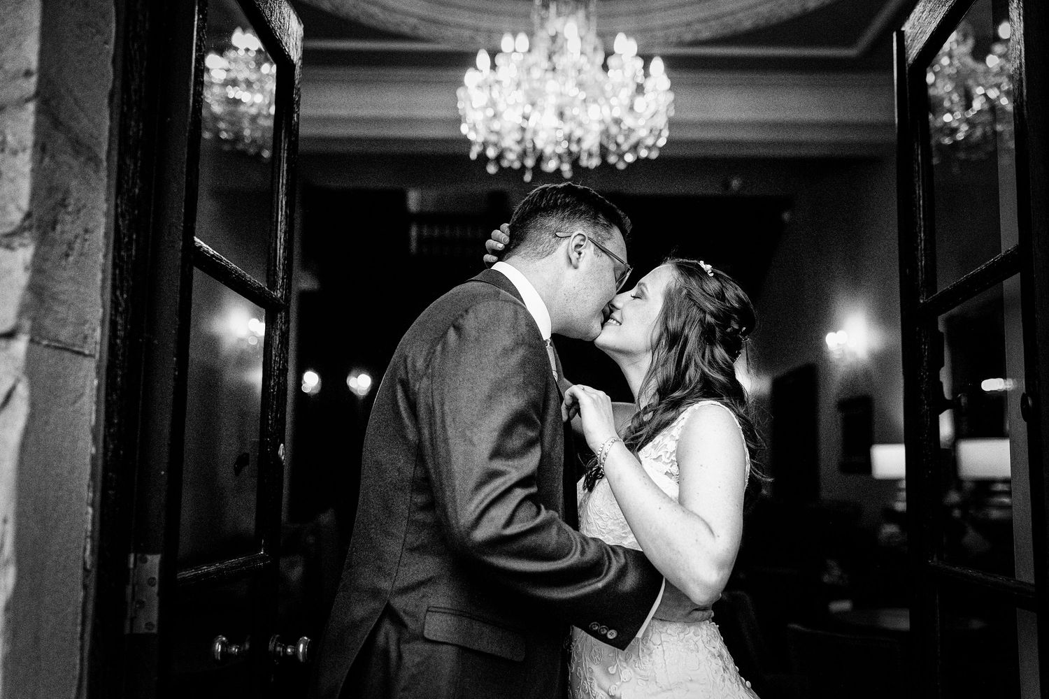 Wedding couple shares tender moments under elegant crystal chandeliers in dark reception venue.