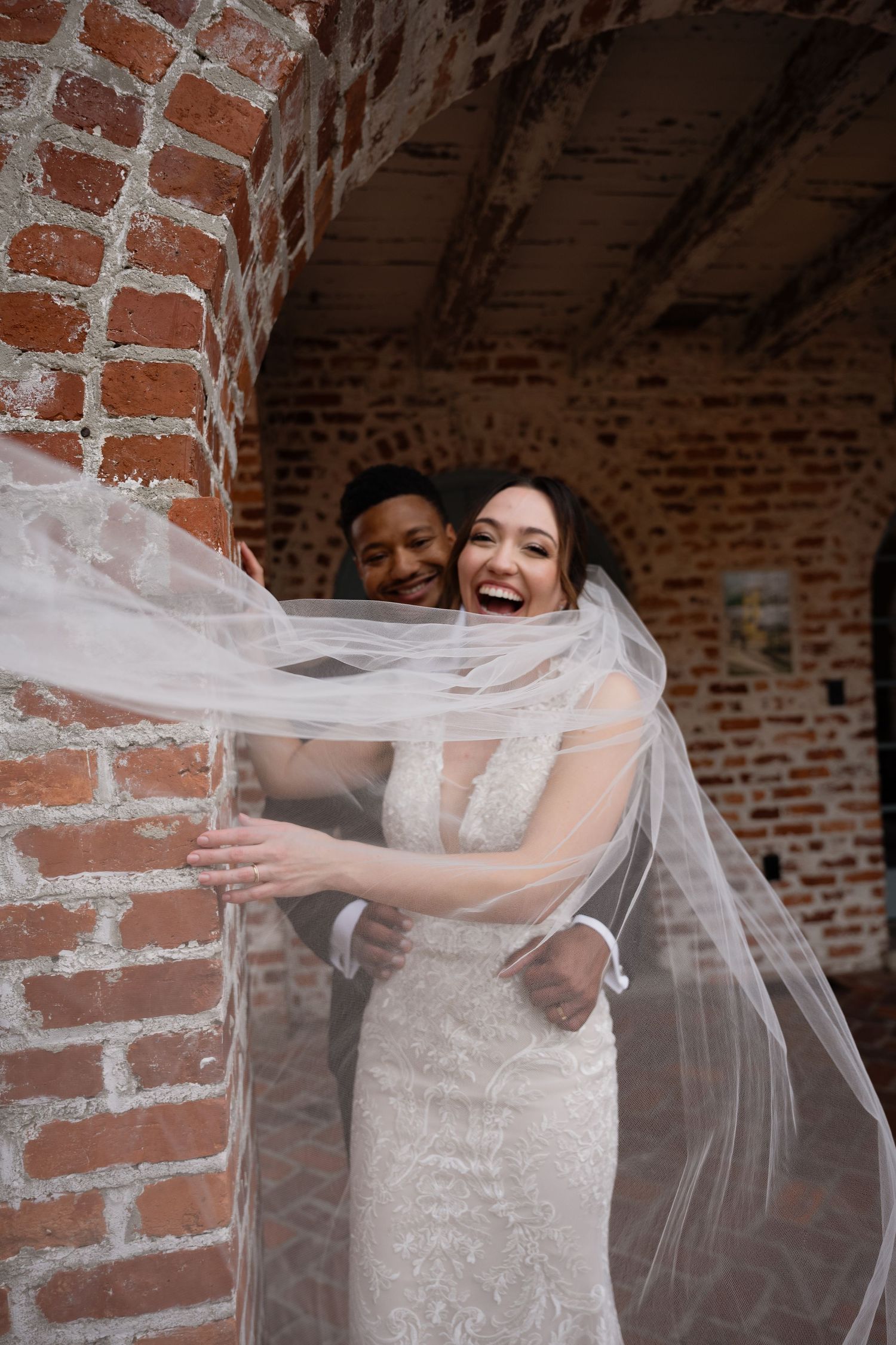 Playful moment between newlyweds with flowing veil against brick wall background.