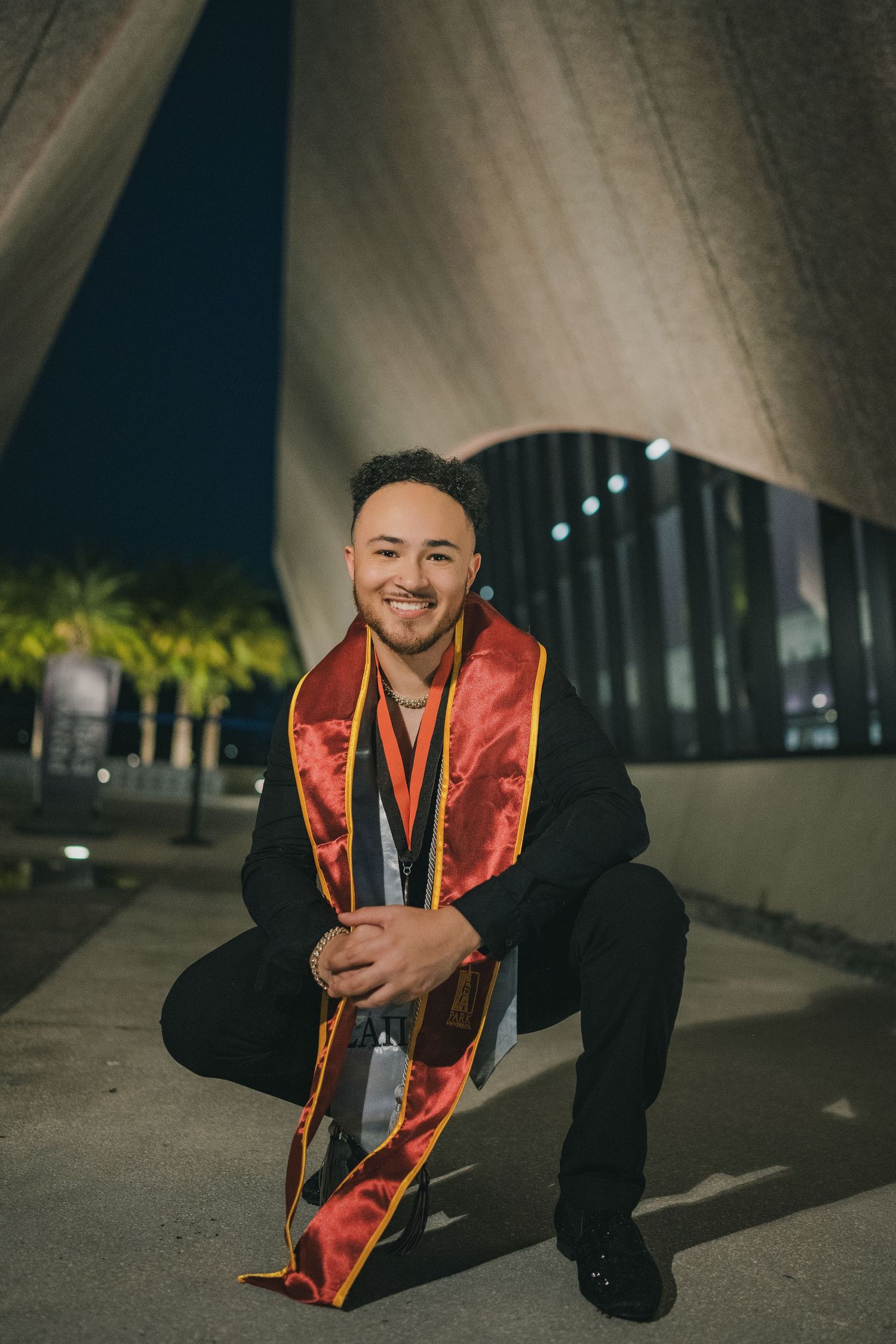 Graduate in red stole and black gown poses at night under architectural lighting.