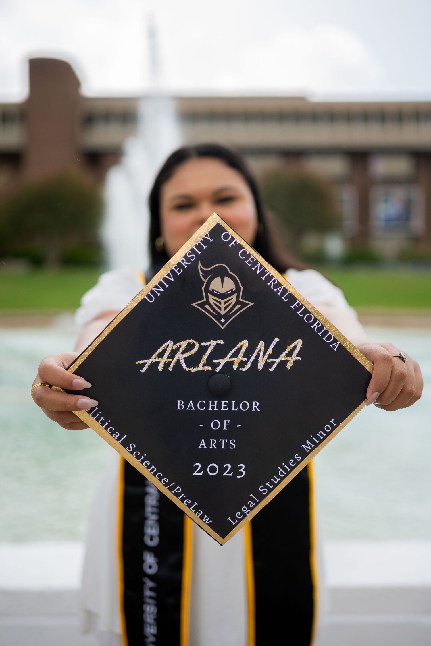 Graduate displaying decorated graduation cap with 'Ariana Bachelor of Arts 2023' against fountain backdrop.