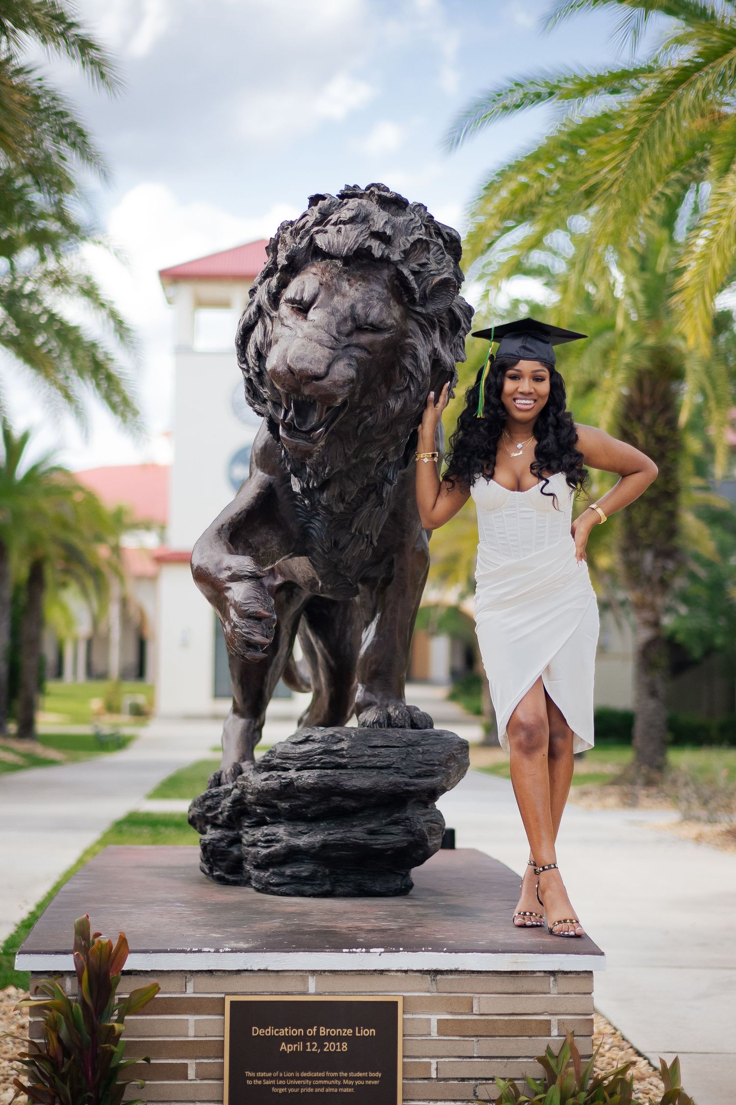 Graduate in white dress poses next to large decorative lion statue on campus walkway.