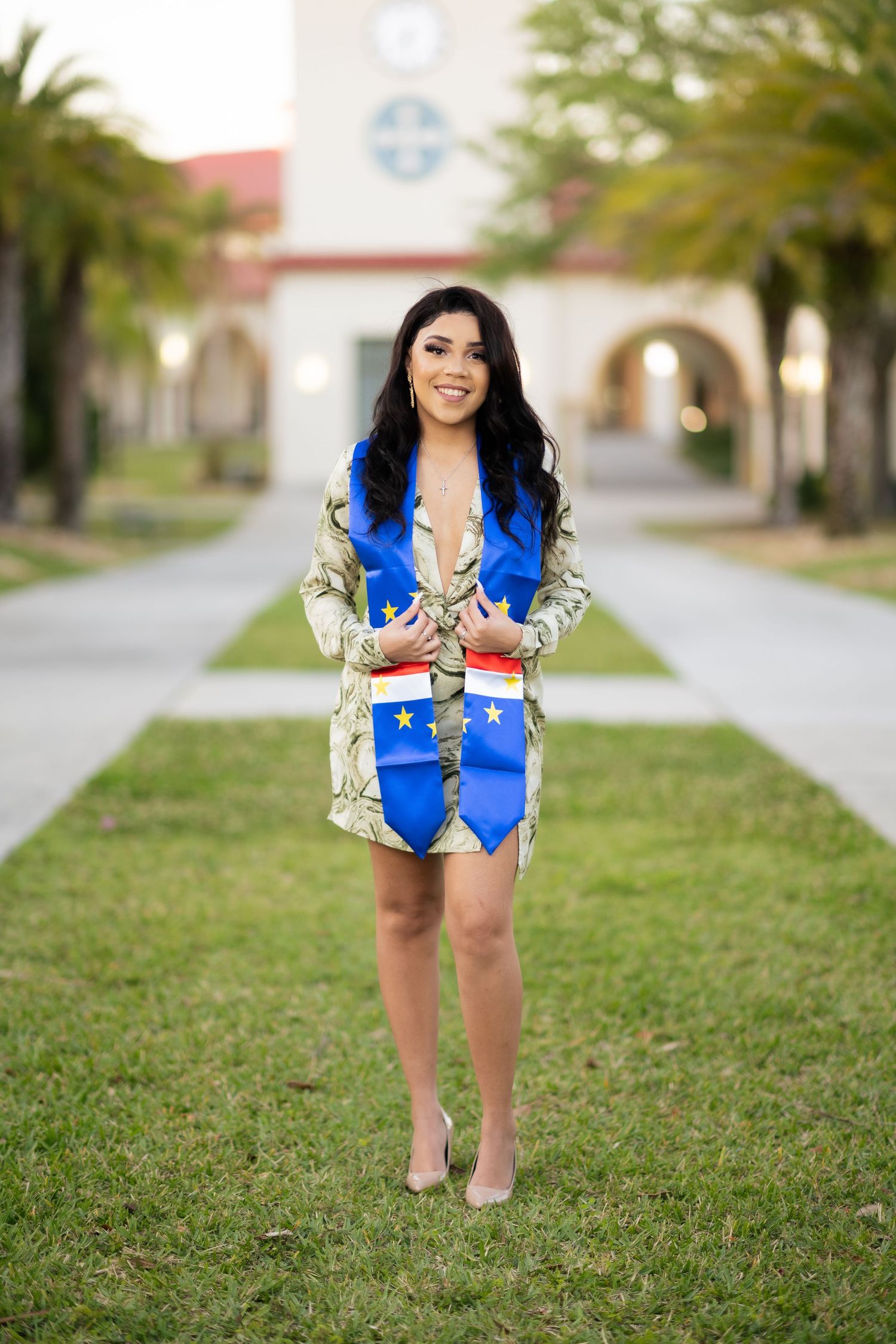Graduate in blue and gold stole strikes a pose on a palm tree-lined pathway near campus buildings.