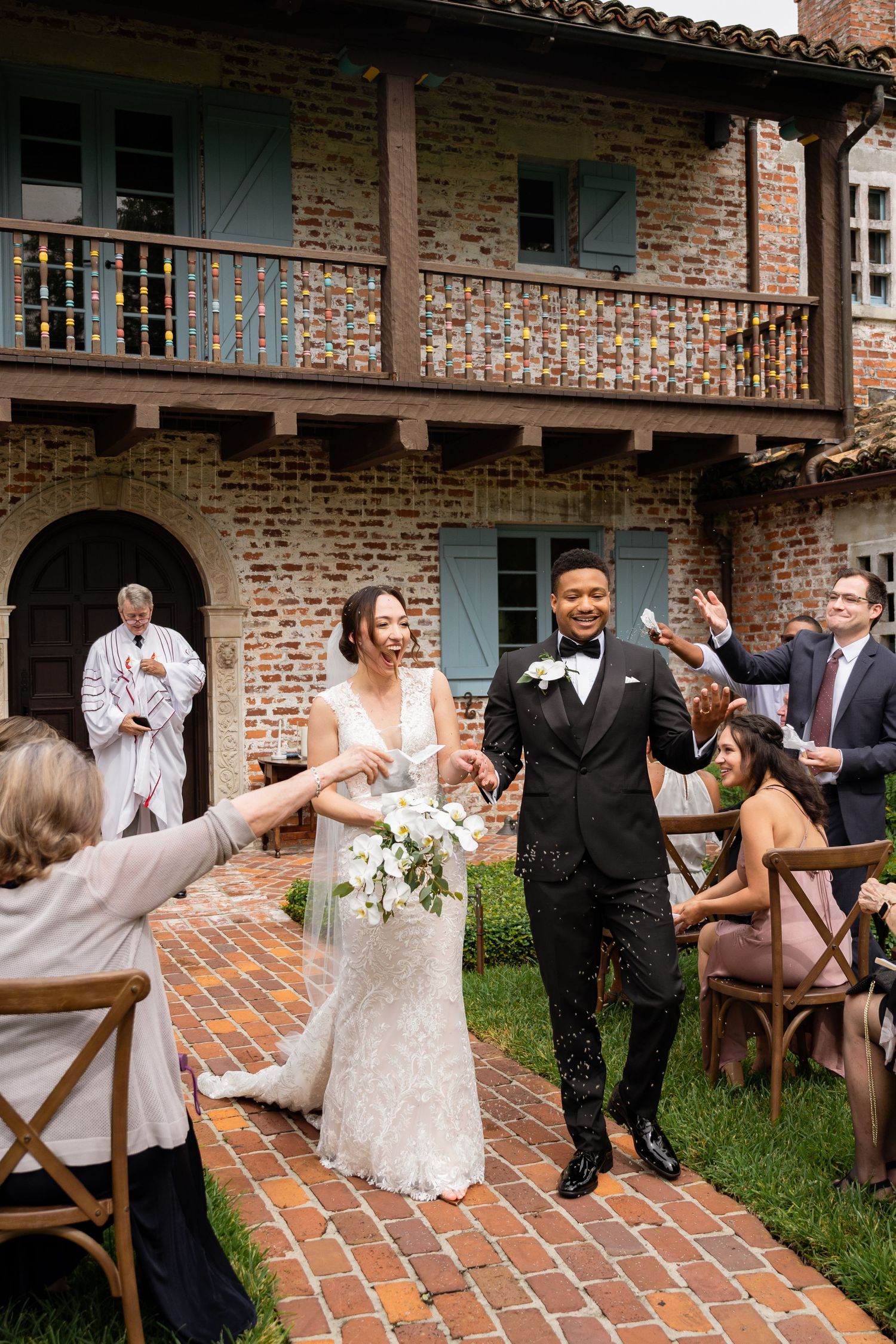 A wedding couple exits their ceremony while guests celebrate and throw confetti at a brick venue.