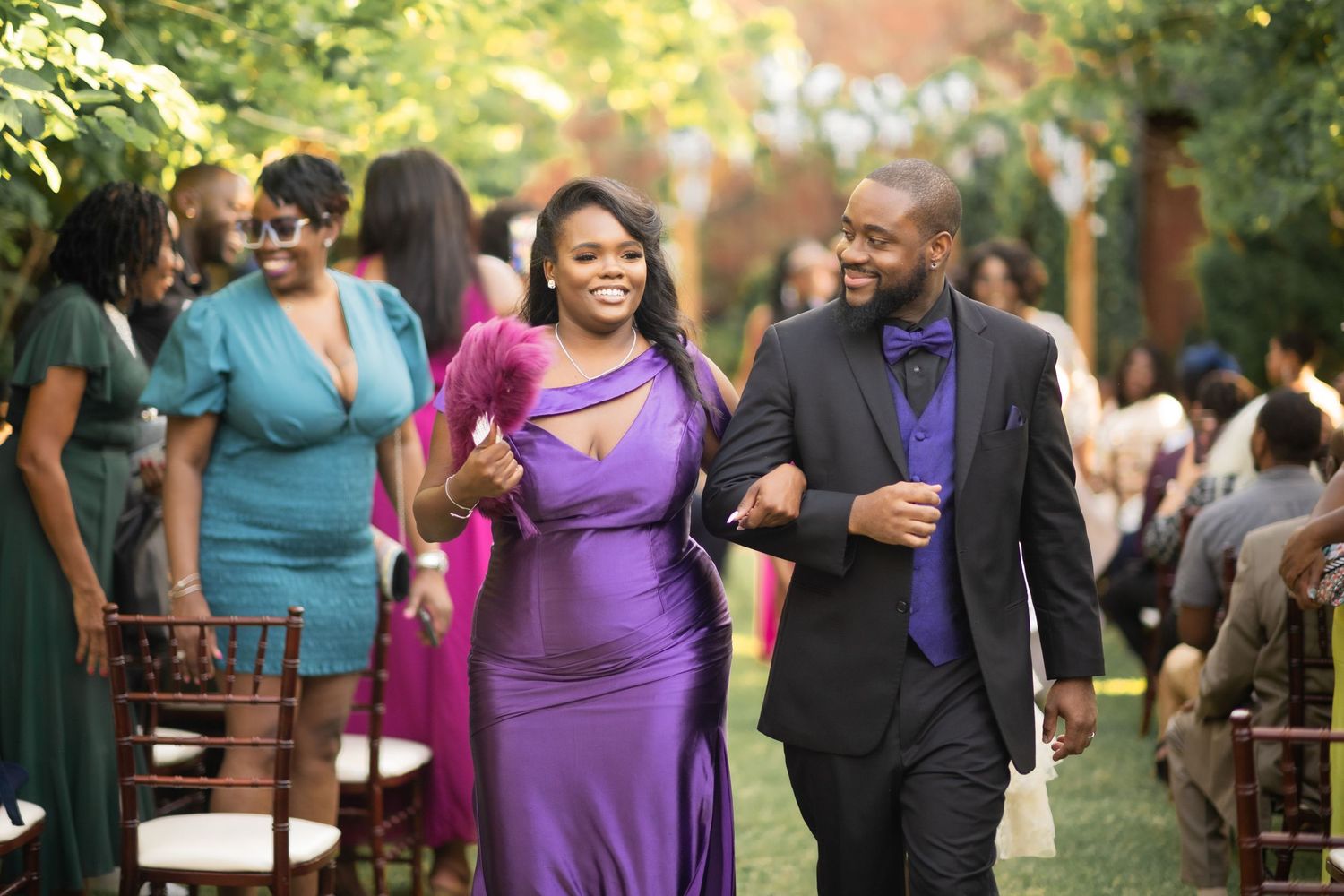 Joyful wedding guests in vibrant purple and blue attire walking down an outdoor aisle at sunset.