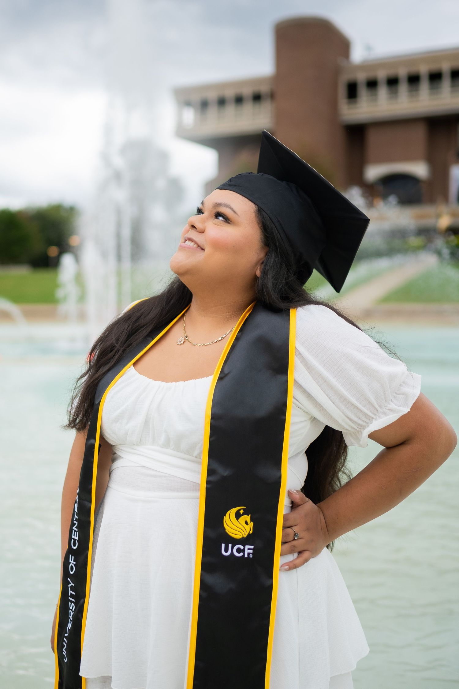 Graduate in white dress and graduation regalia poses confidently by campus fountain.