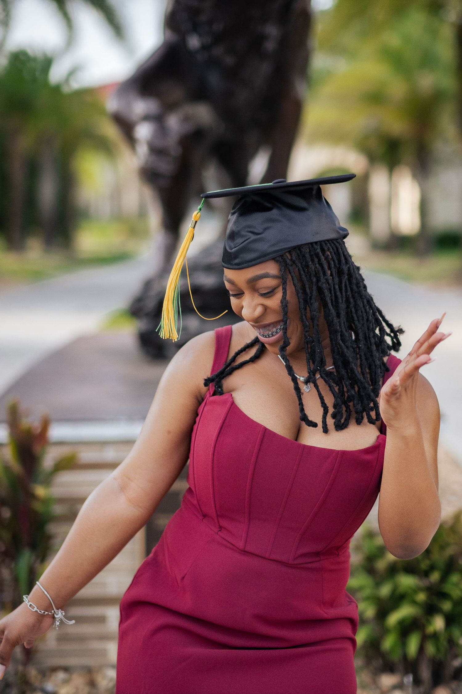Graduate in burgundy dress and black cap poses playfully outdoors.