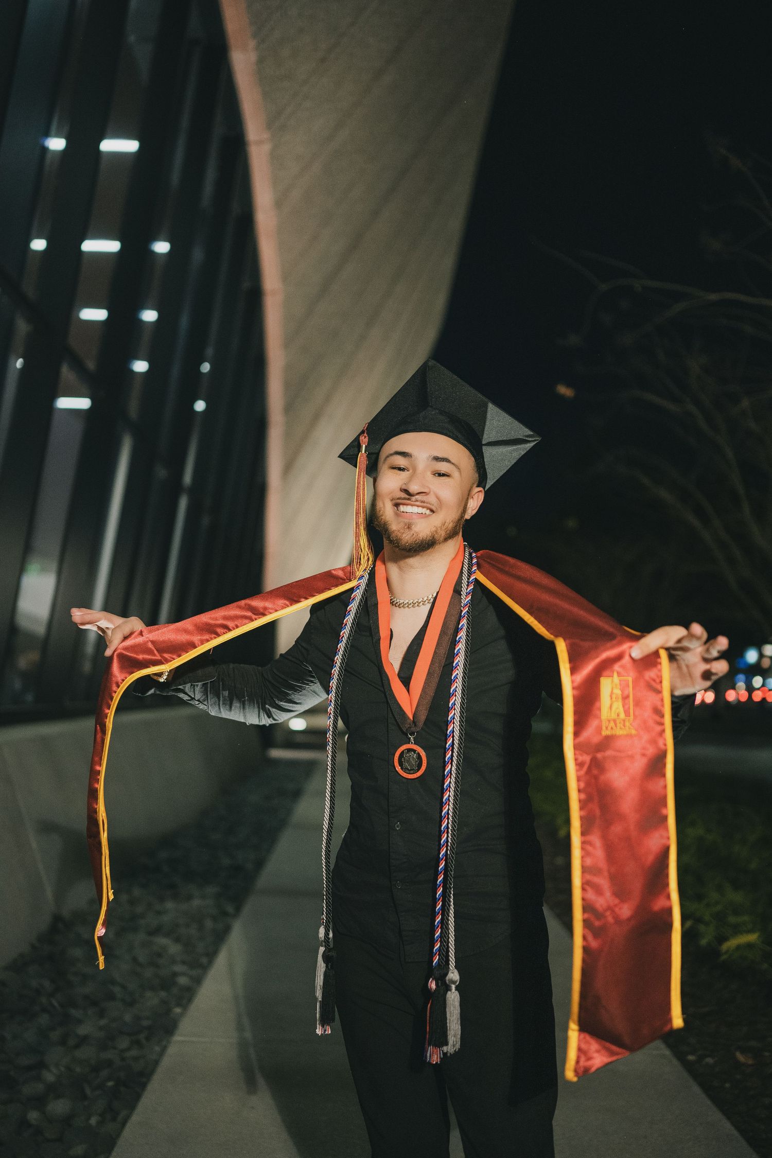 Graduate spreads arms while wearing red stole and black graduation attire at night.