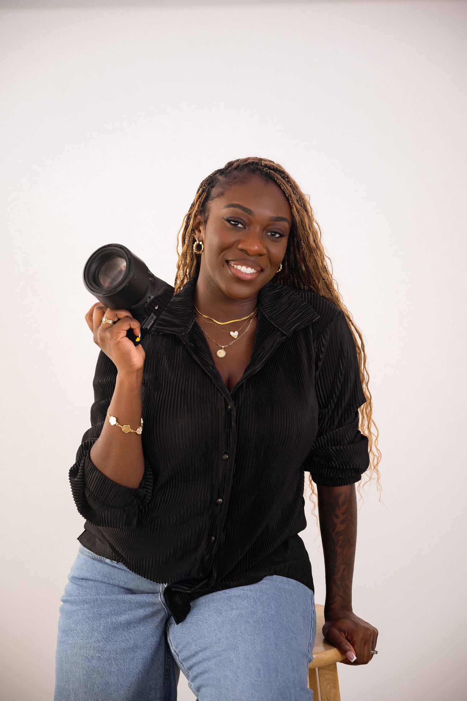 Smiling woman holding a camera in a black shirt and jeans, posing against a white background.