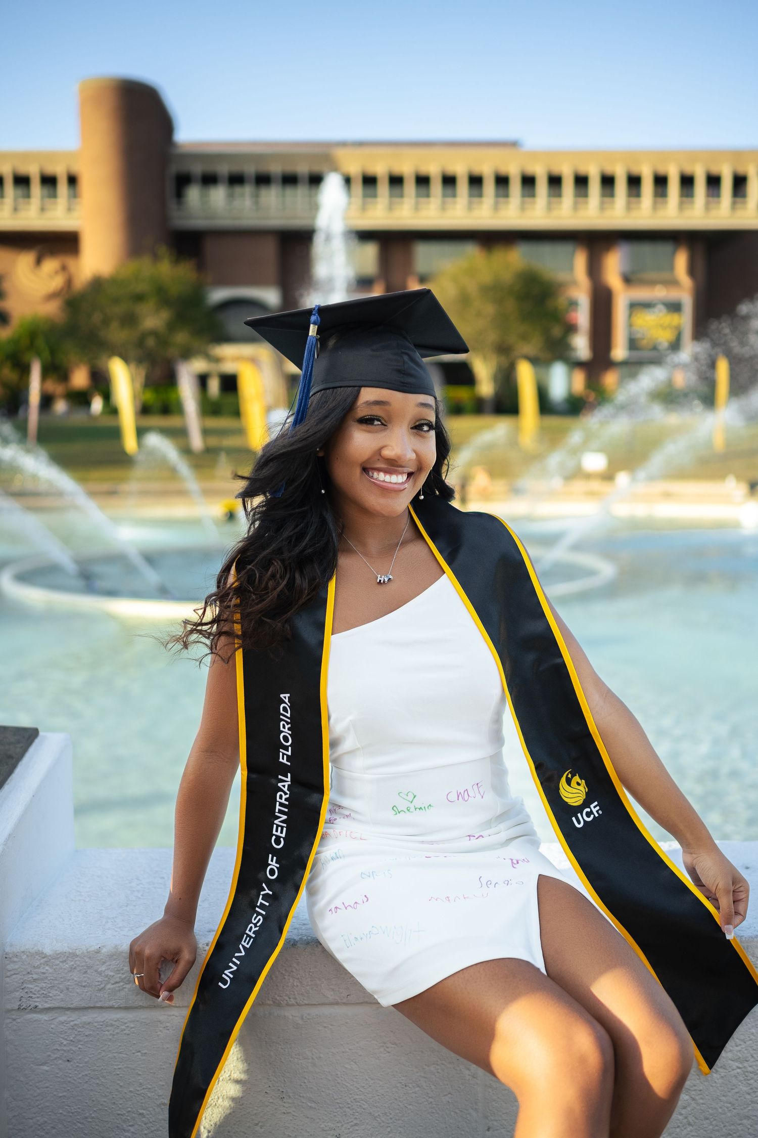 Graduate in white dress and black graduation gown poses by fountain on campus.