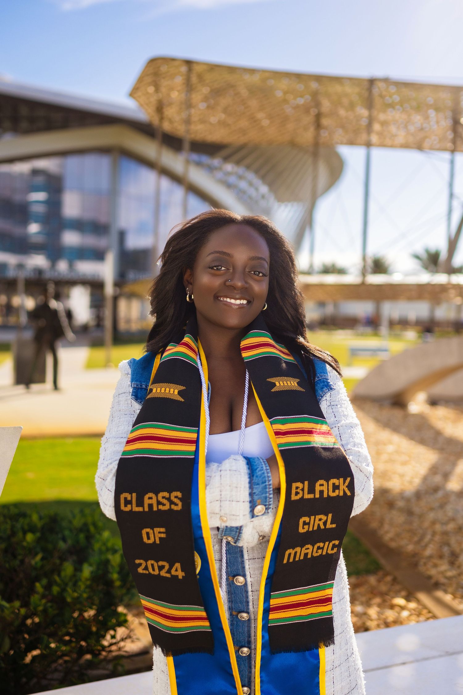 Graduate wearing Class of 2024 stole smiles brightly outside modern campus building.