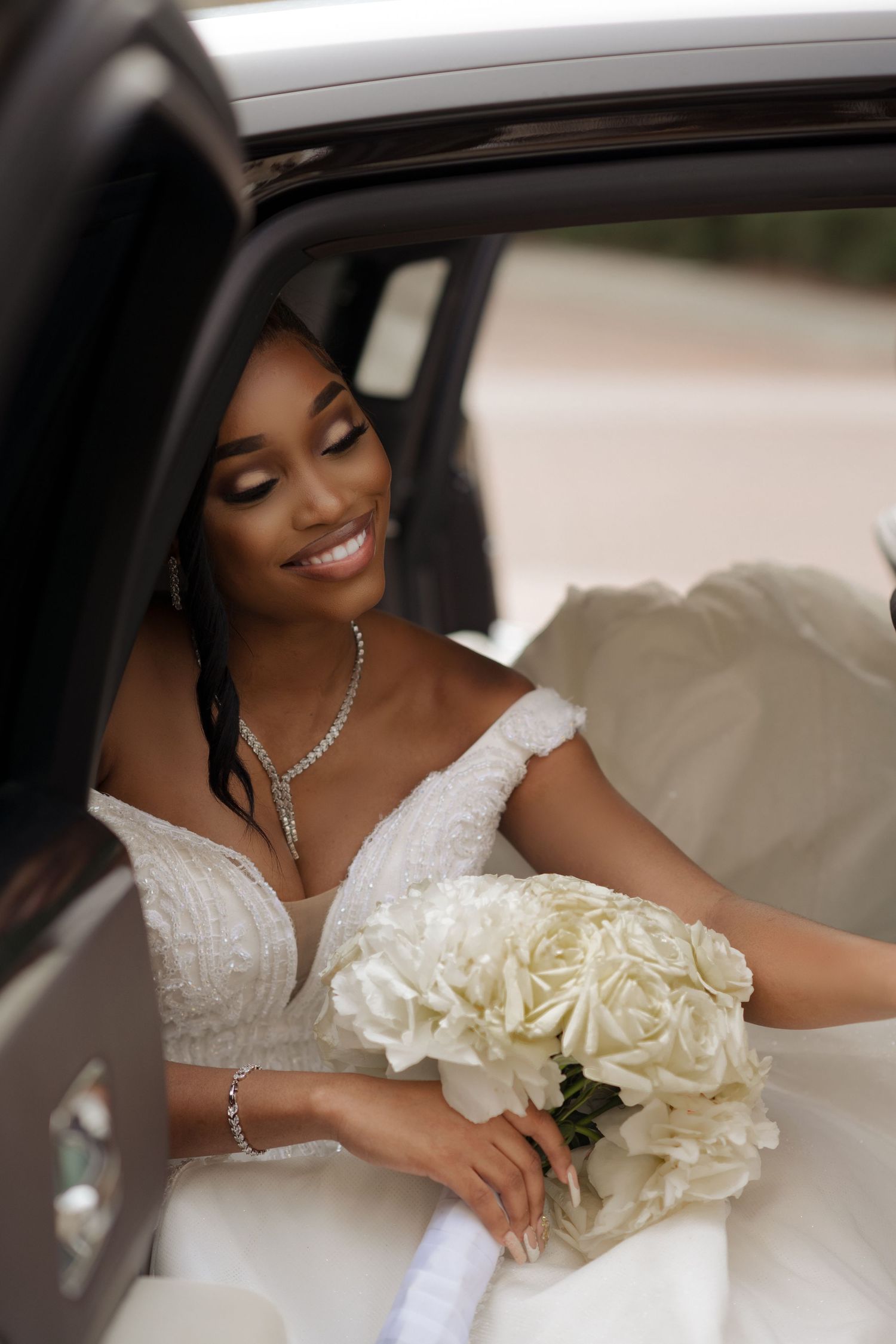 A bride in a white dress holding a cream rose bouquet smiles while seated in her wedding car.