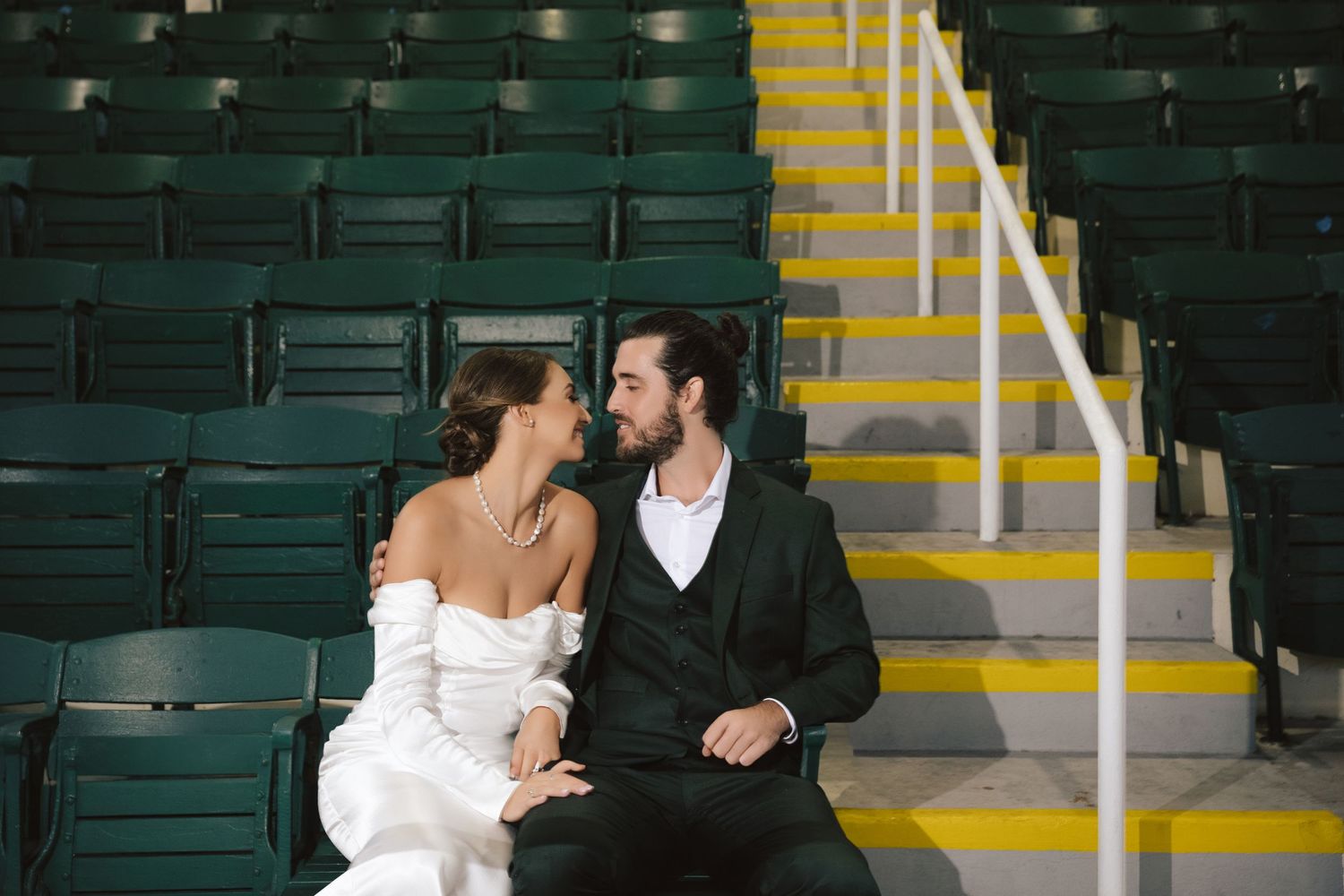 A romantic couple in wedding attire sits intimately on green stadium seats.