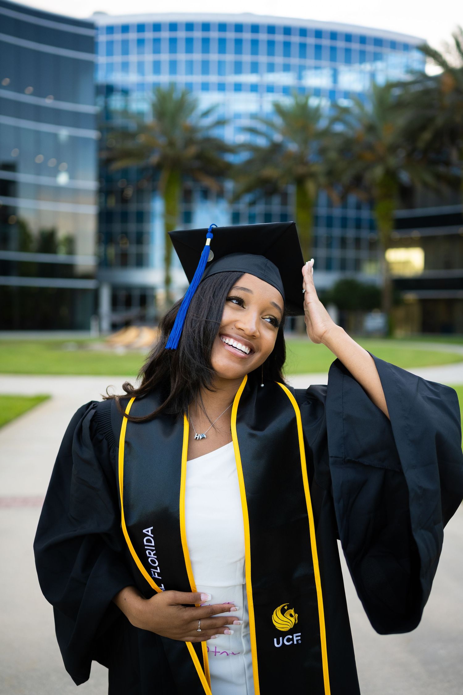Graduate in cap and gown posing outside modern glass building with palm trees.