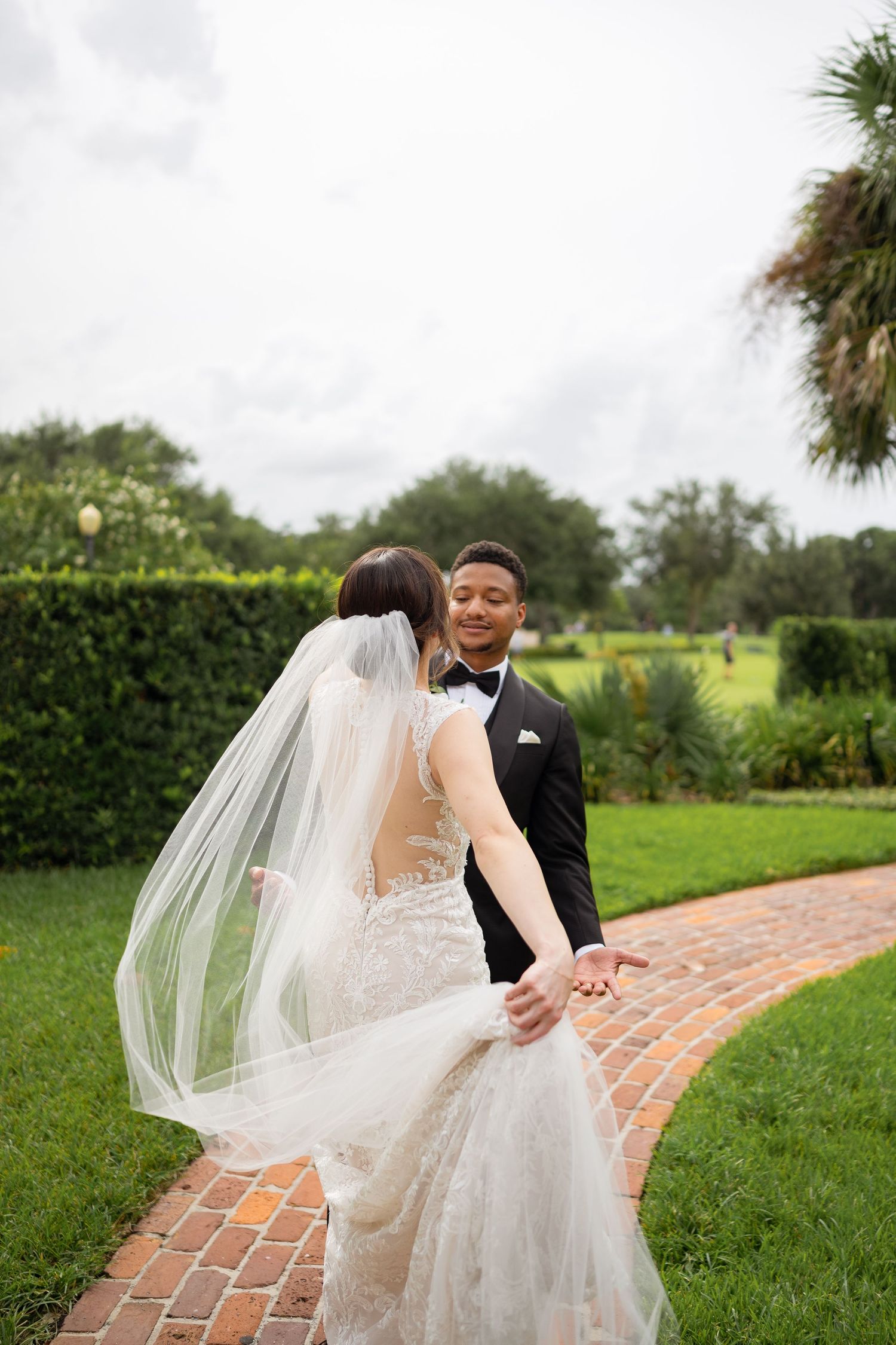 Elegant bride in lace gown with flowing veil on brick garden pathway.