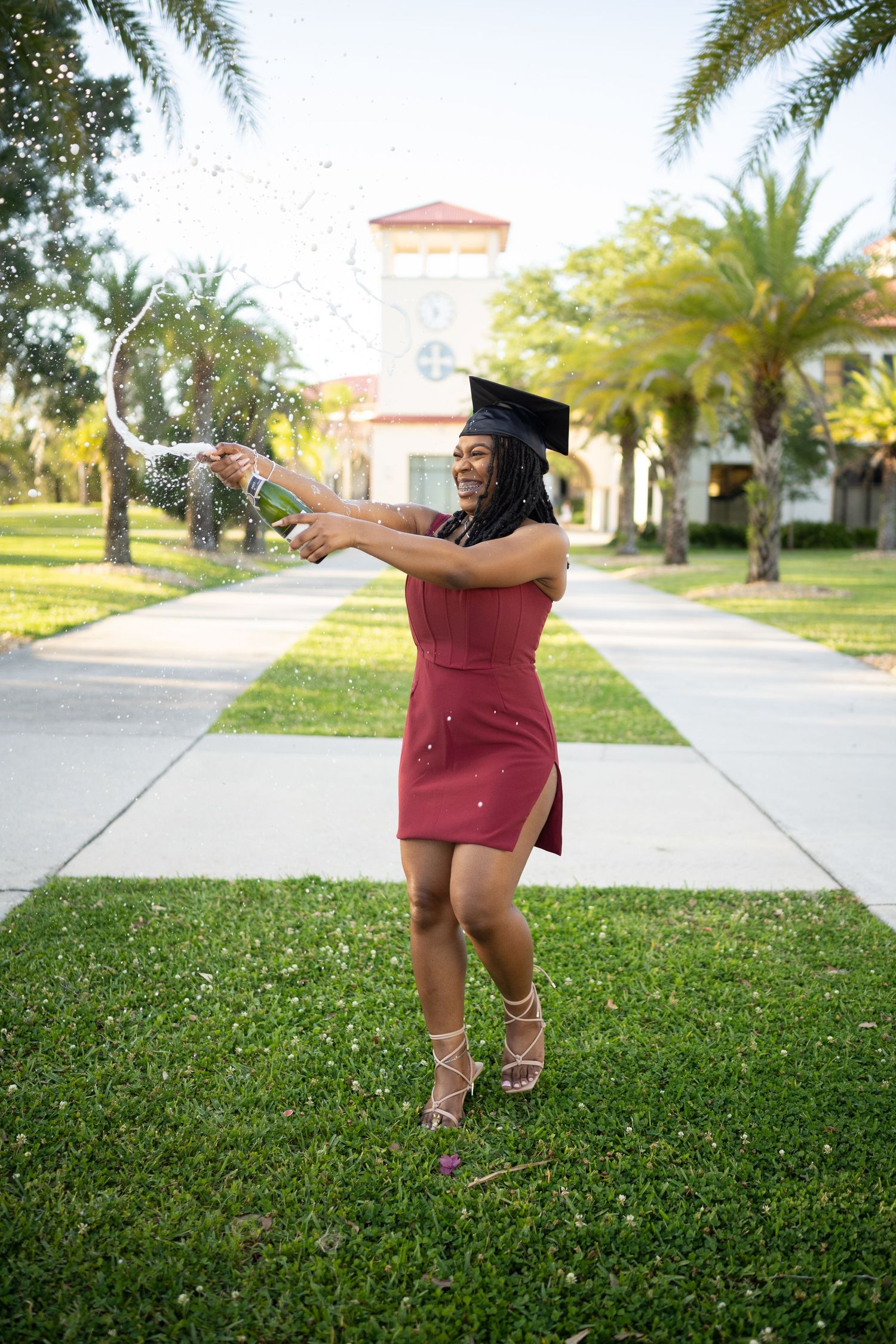 Graduate in burgundy dress throws glitter in sunny campus courtyard.