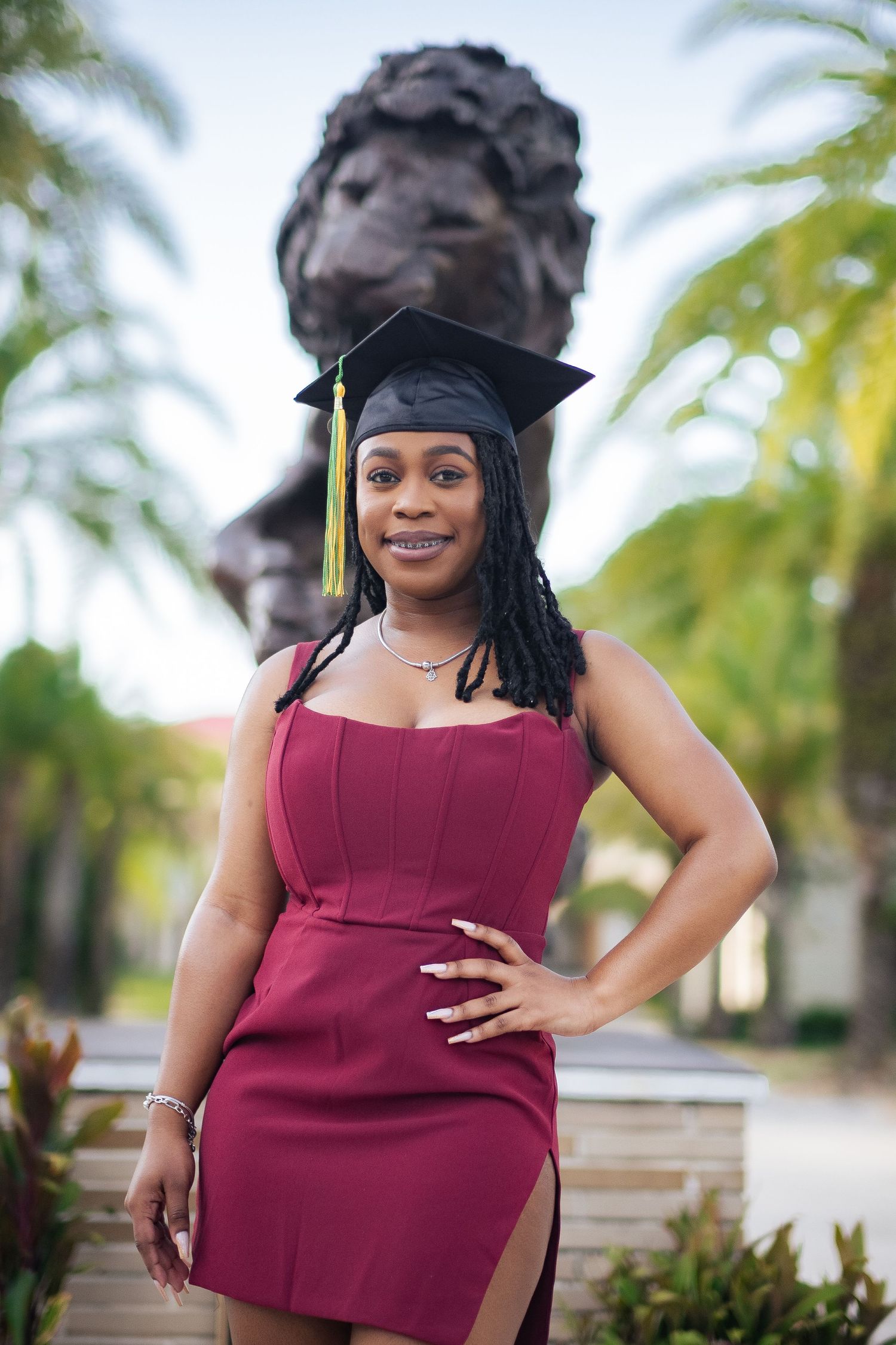 Graduate in burgundy dress poses with statue and palm trees in background.