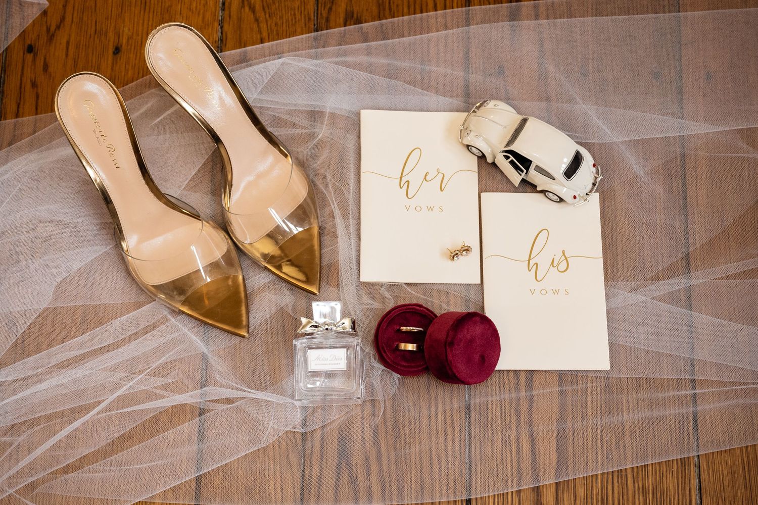 Wedding details flatlay with gold heels, perfume bottle, ring box and vows cards on tulle fabric.