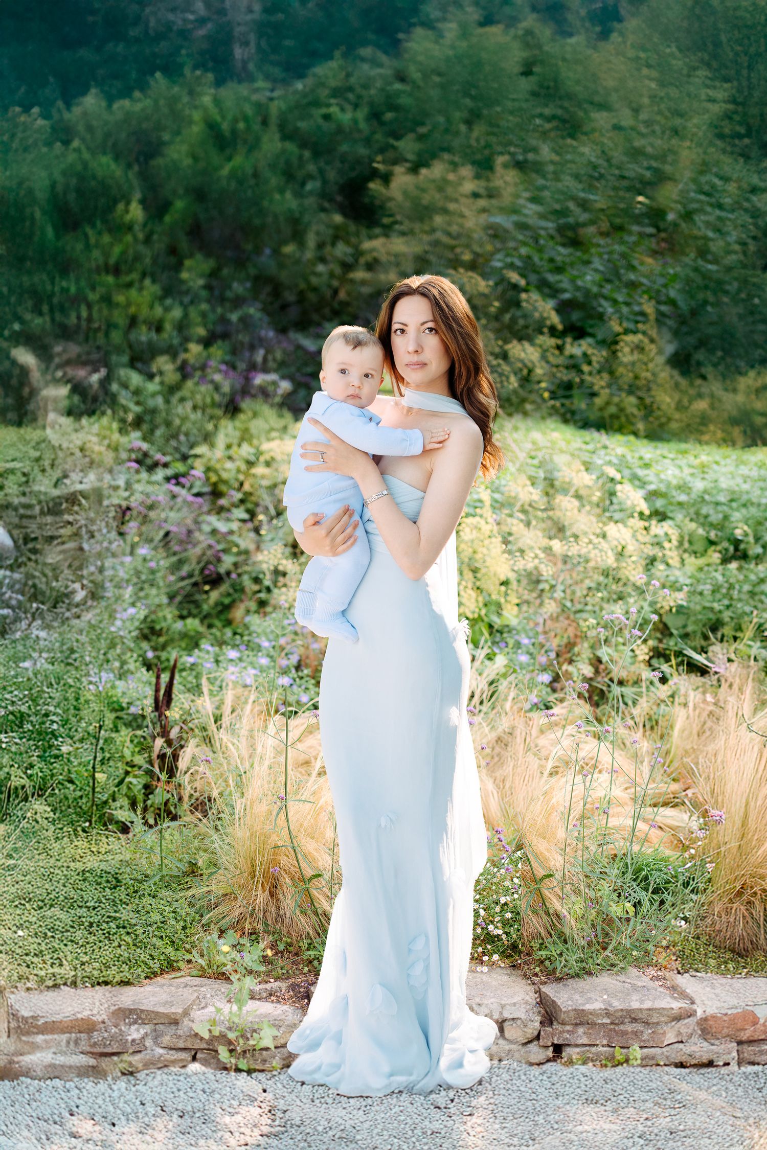 A woman in an elegant light blue gown holding a baby stands in a garden setting with greenery in the background.