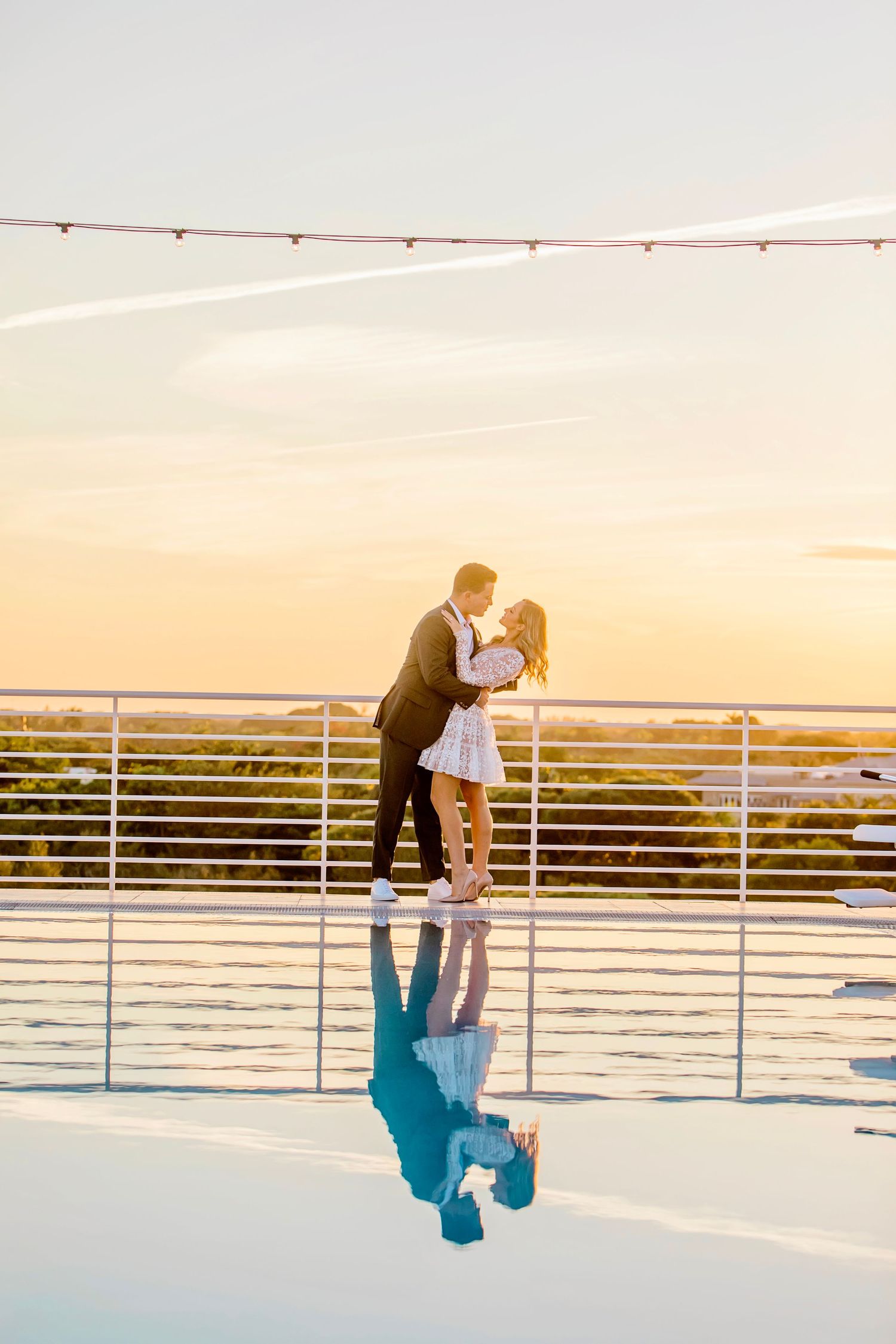 Couple sharing romantic moment at sunset with reflection on glossy surface and string lights above.