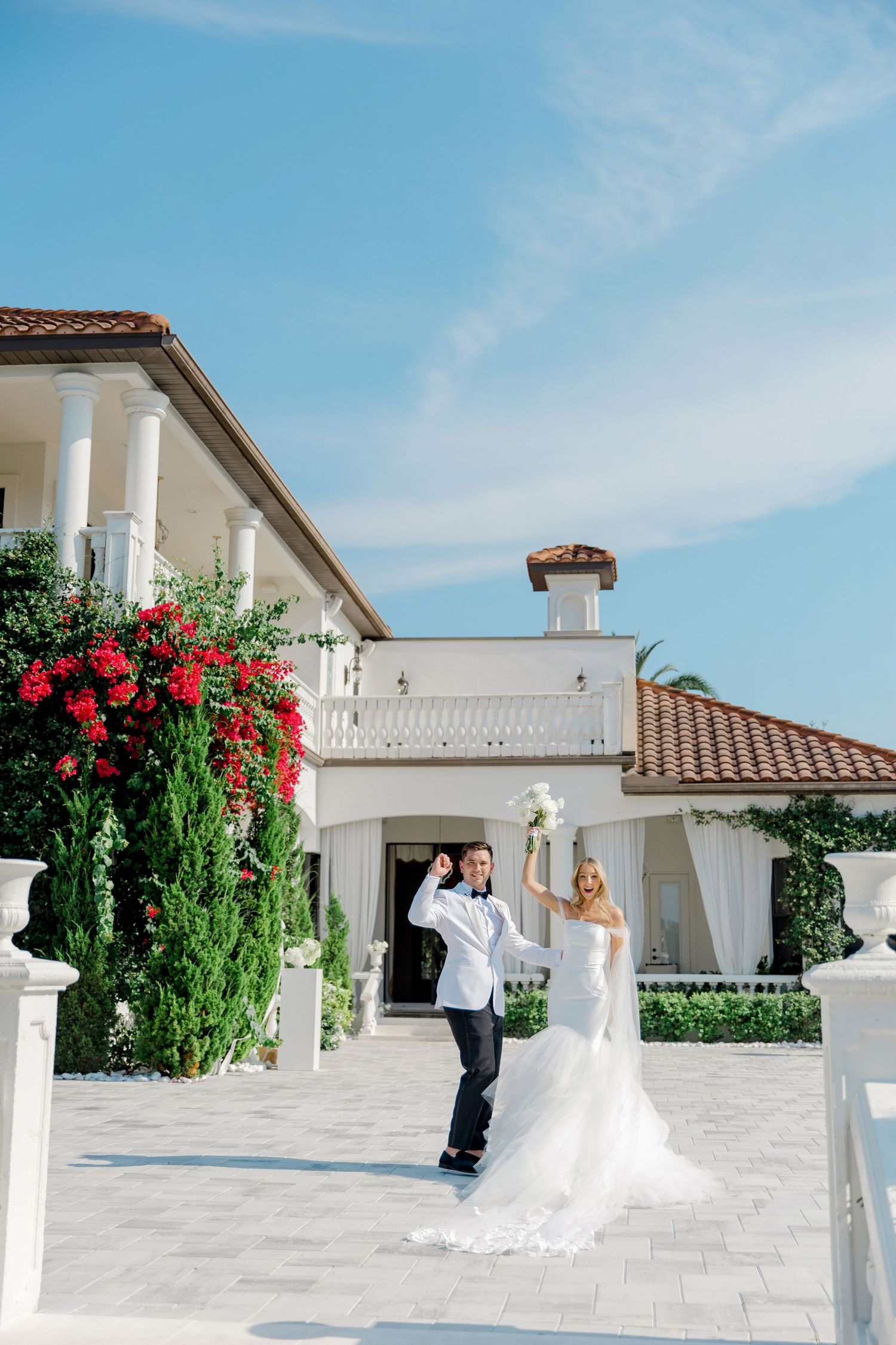 Newlyweds celebrating outside elegant white Mediterranean-style villa with red flowers.