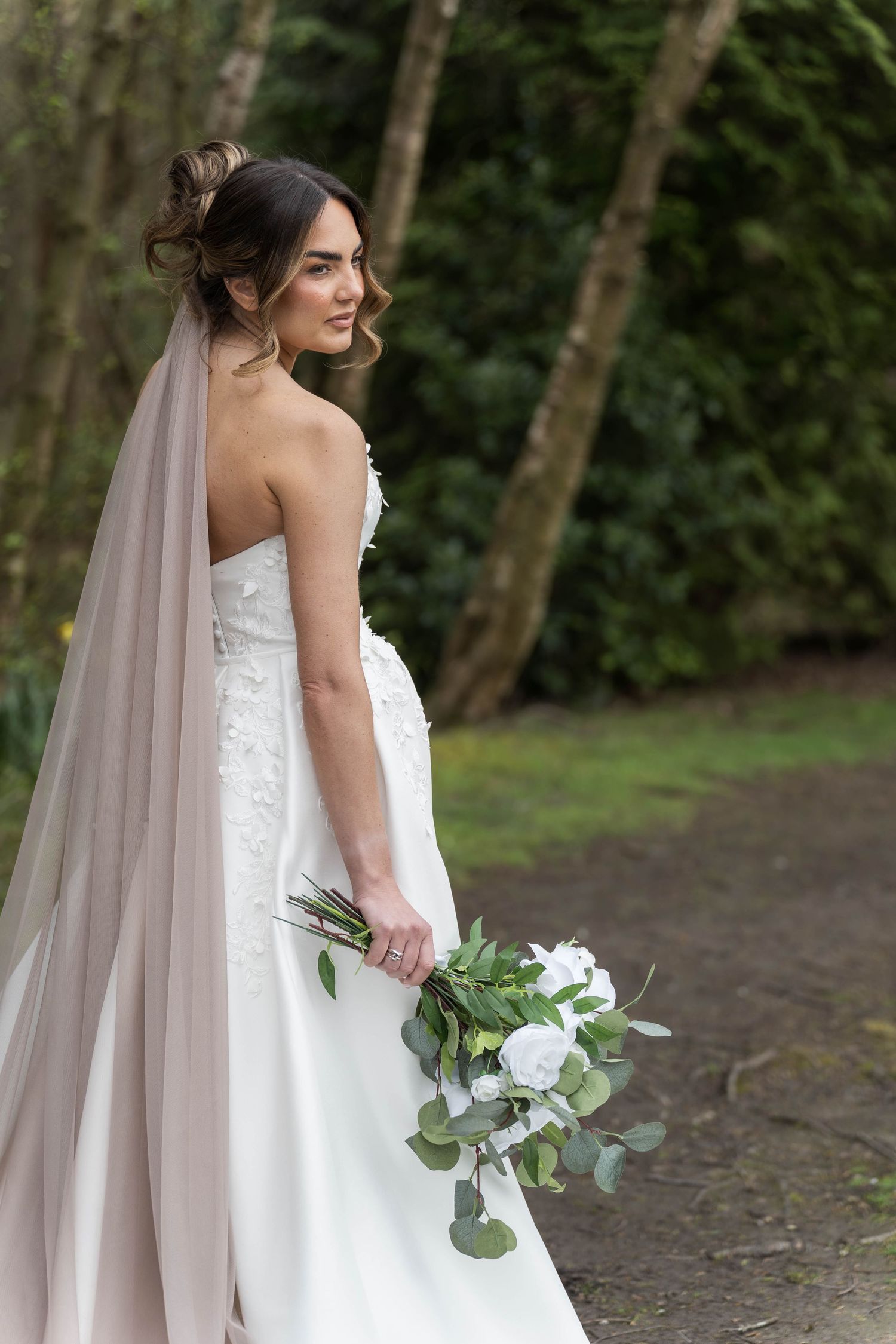 Elegant bride in strapless lace gown with blush veil holding white floral bouquet outdoors.
