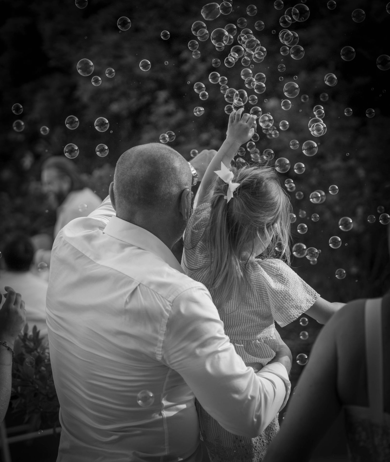 Black and white photo of a bald man holding a young girl reaching for floating bubbles outdoors.