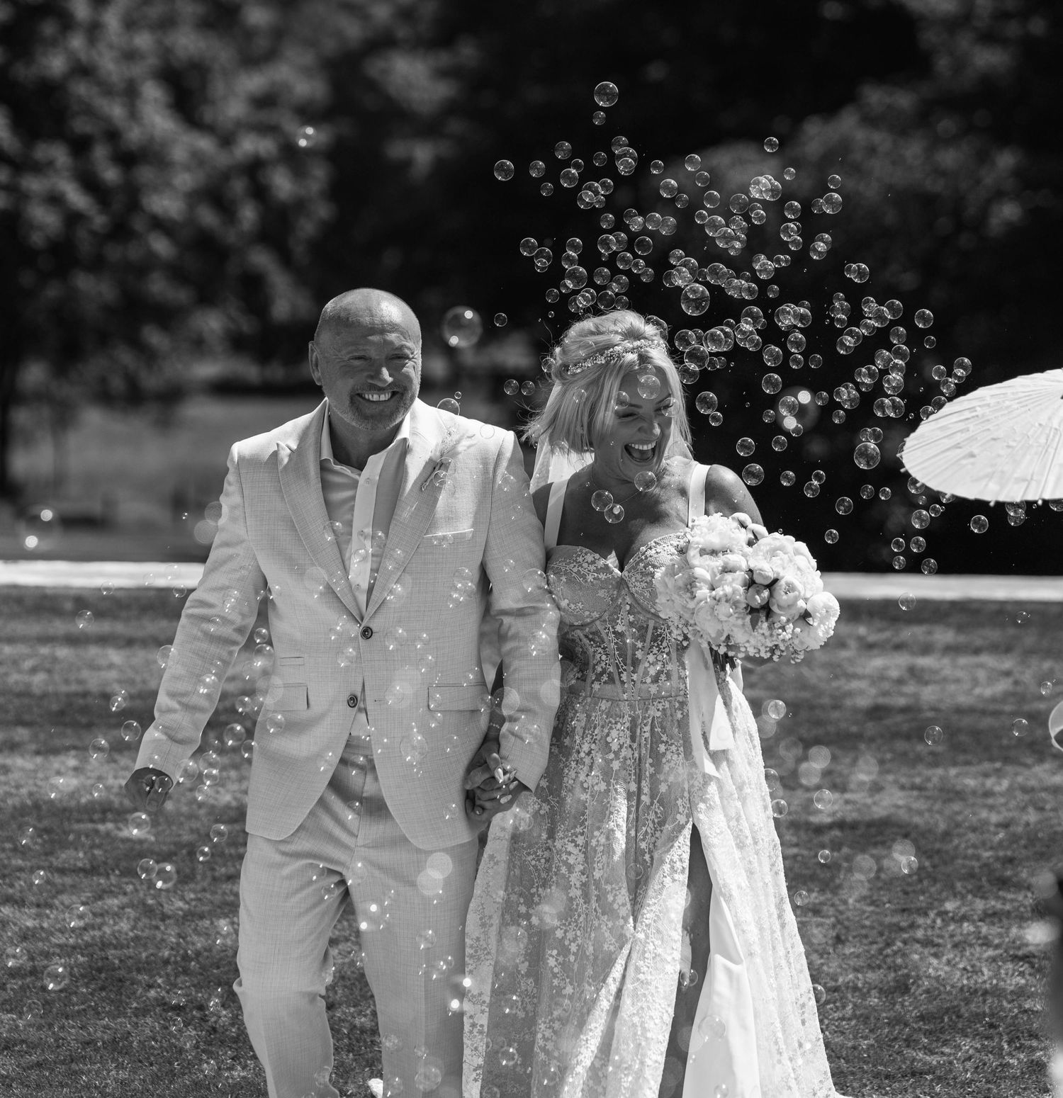 Black and white photo of a joyful bride and groom walking through bubbles at their outdoor wedding ceremony.