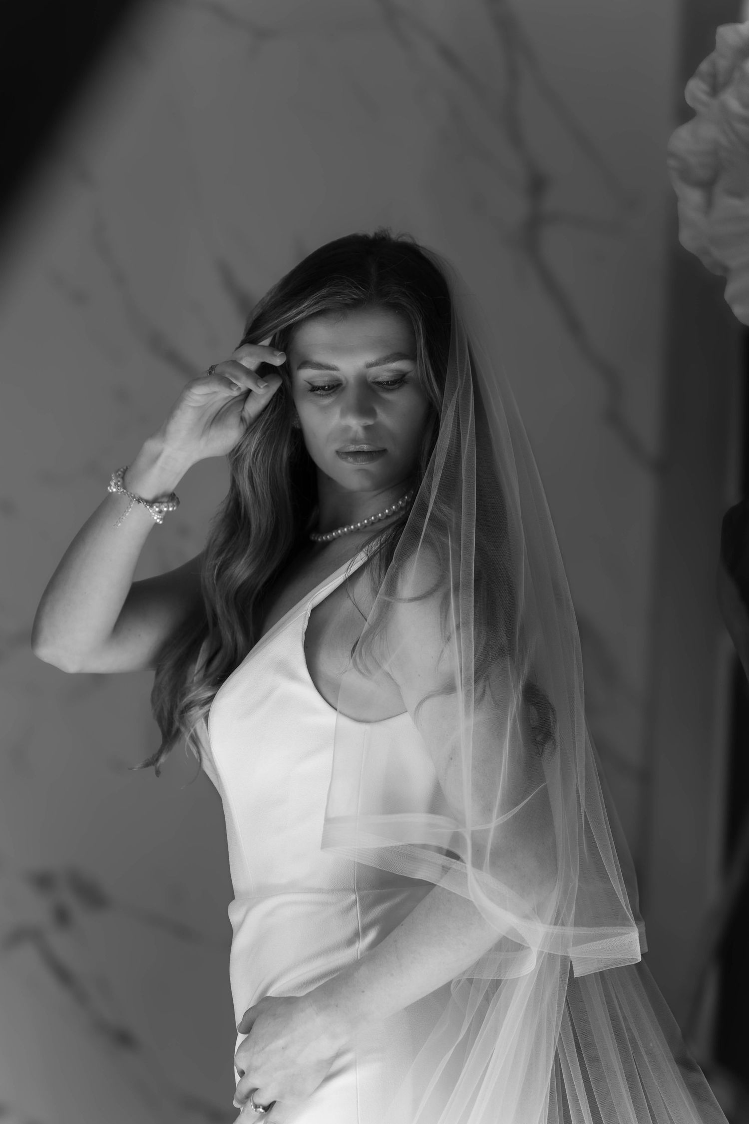 Black and white bridal portrait of a woman in a wedding dress and veil, adjusting her hair with jewelry.