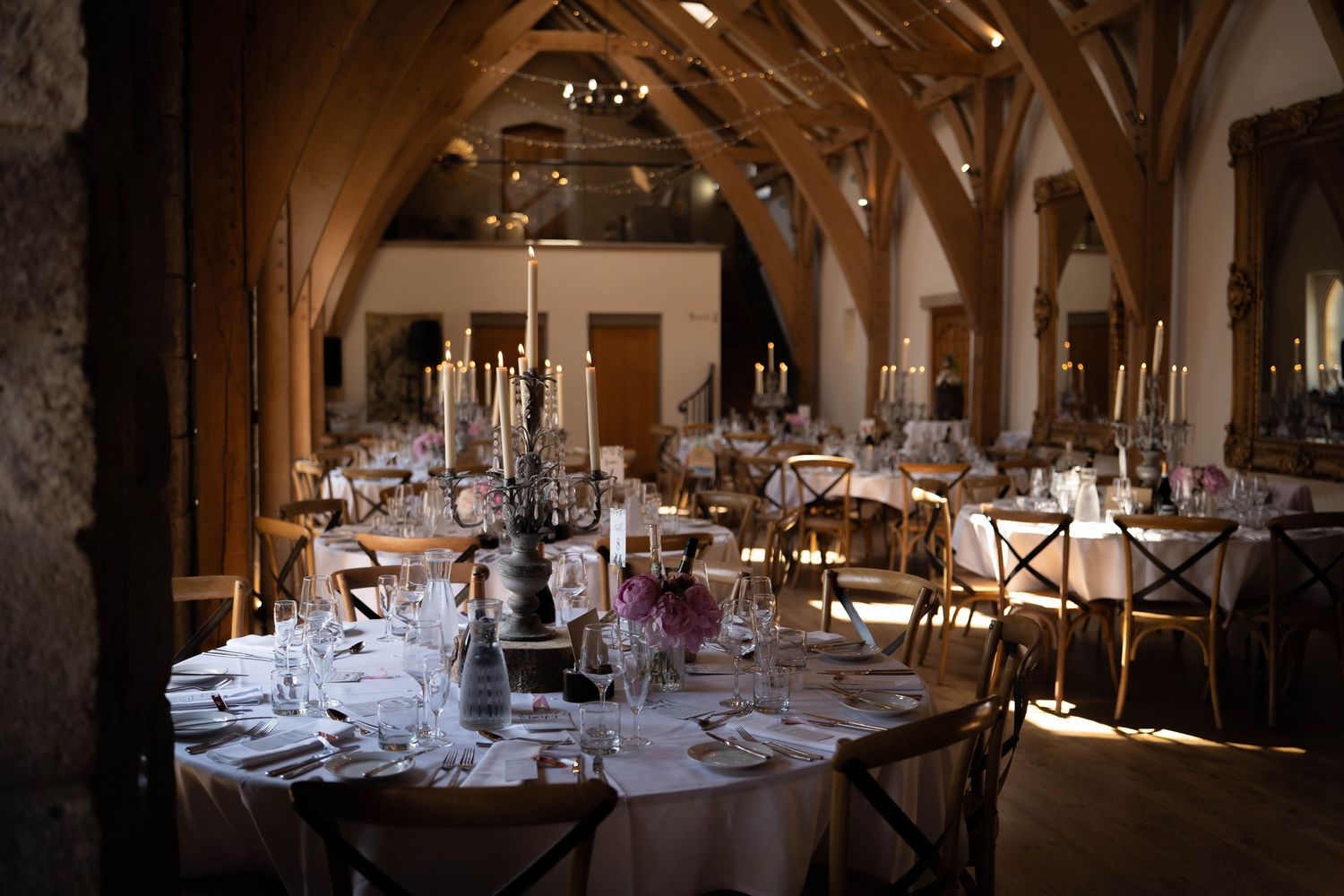 Elegant barn wedding reception with round tables, candlelit centerpieces, and rustic wooden arch beams in a historic venue.