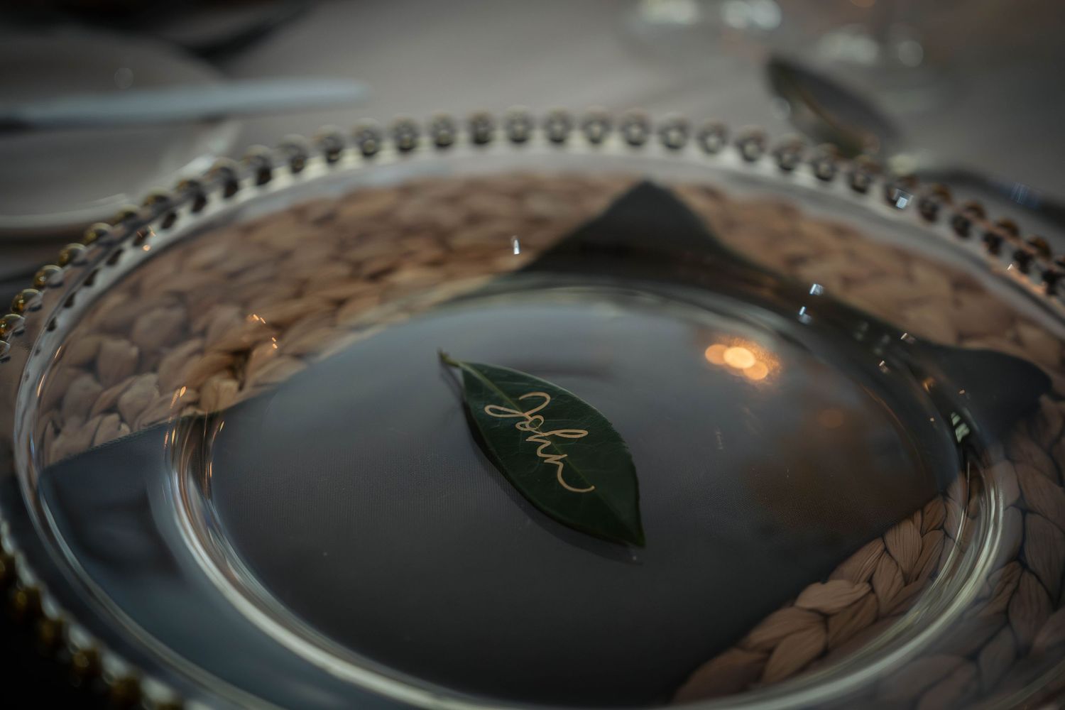 Elegant place setting with a silver charger plate, woven placemat, and a green leaf name card for a formal event.