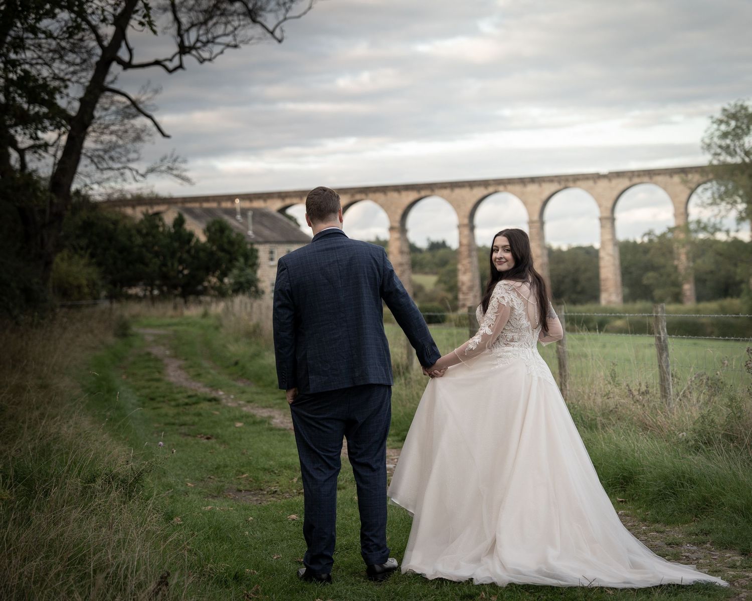 Newlyweds stand together against a scenic landscape with an old stone viaduct in the distance.