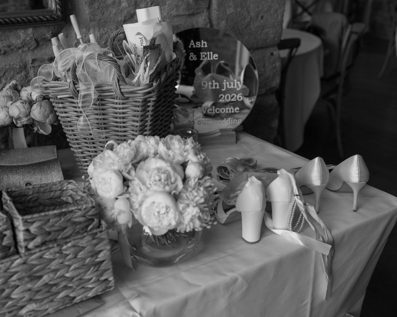 Black and white wedding table with floral arrangement, wicker baskets, bridal heels, and personalized welcome sign for Ash and Elle 2026.