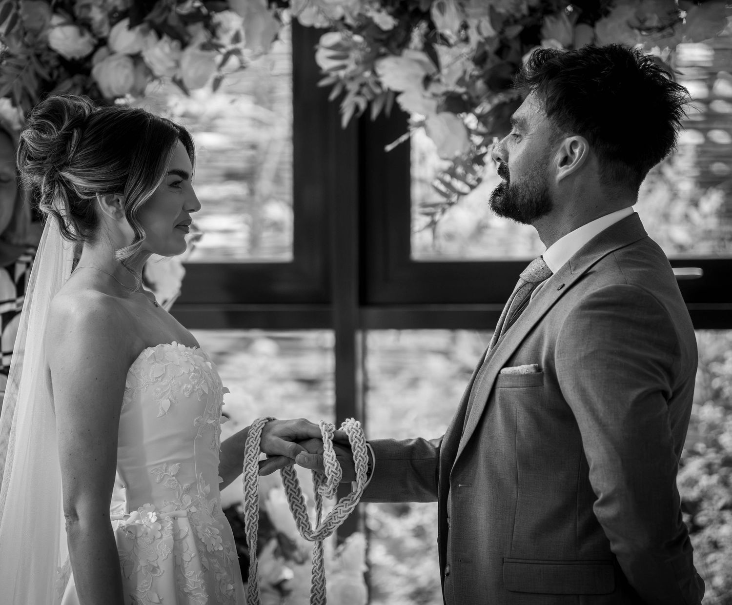 Black and white wedding photo of bride and groom holding hands at altar with cross and floral backdrop.