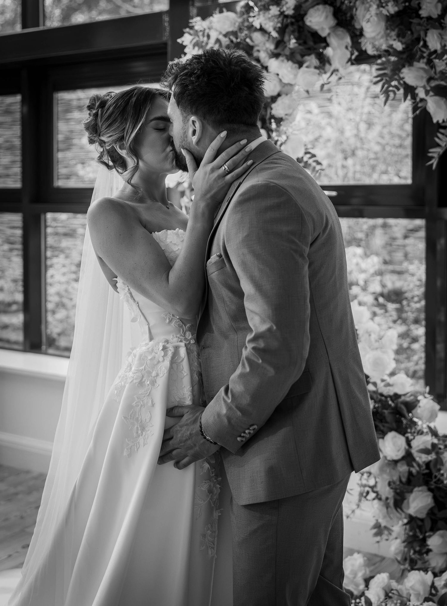 Black and white photo of a bride and groom sharing their first kiss at the altar adorned with floral arrangements.