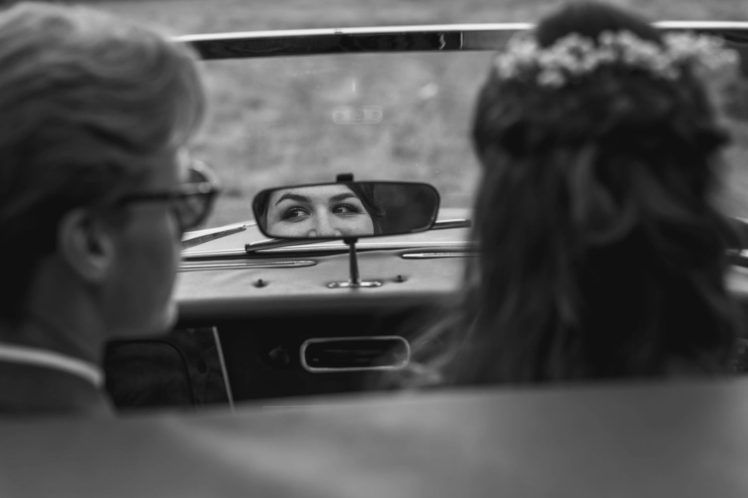 Black and white wedding photo of bride reflected in car rearview mirror, with groom in foreground.