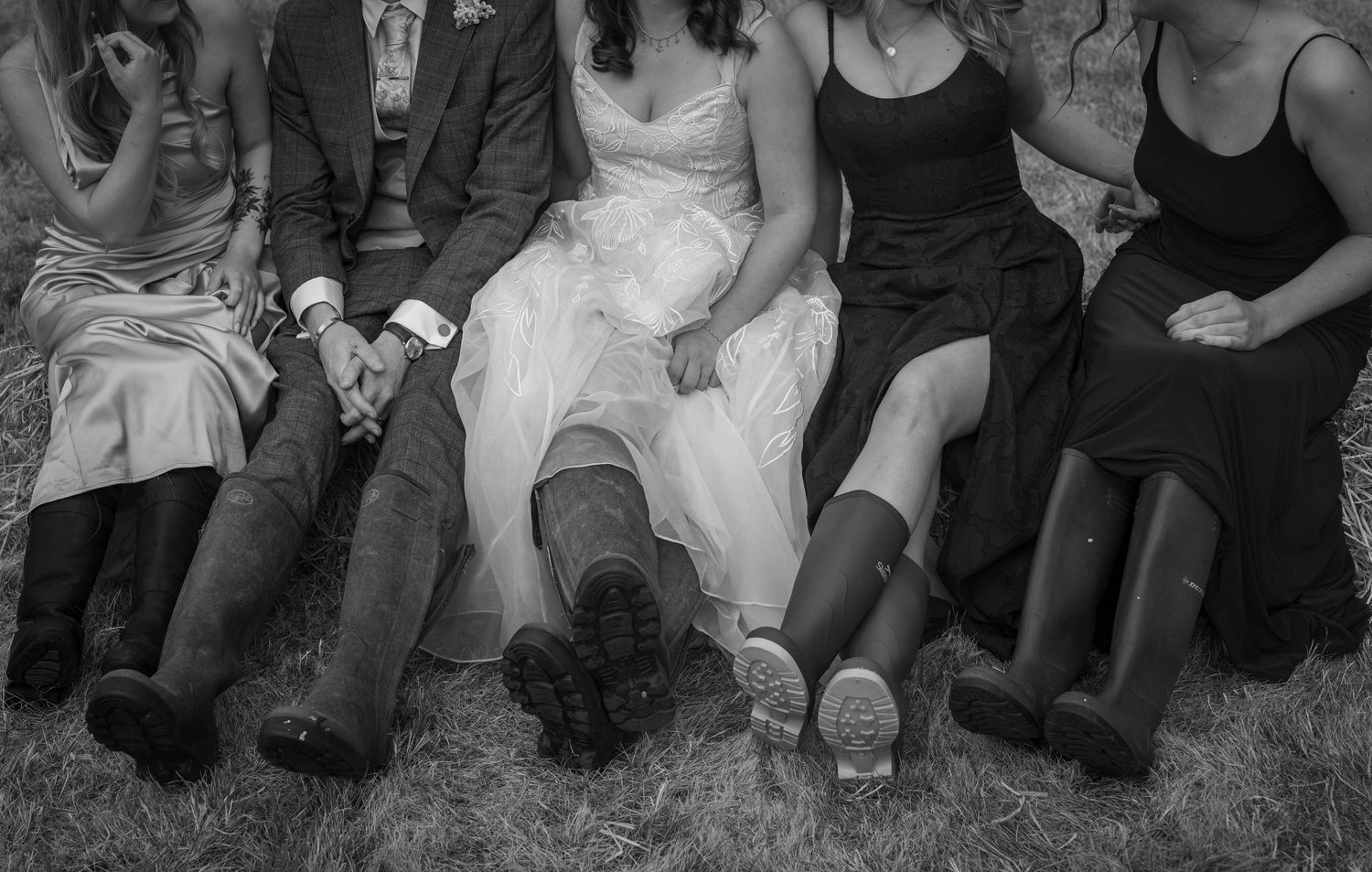 Black and white wedding photo of bride, groom, and bridesmaids sitting outdoors wearing rain boots.