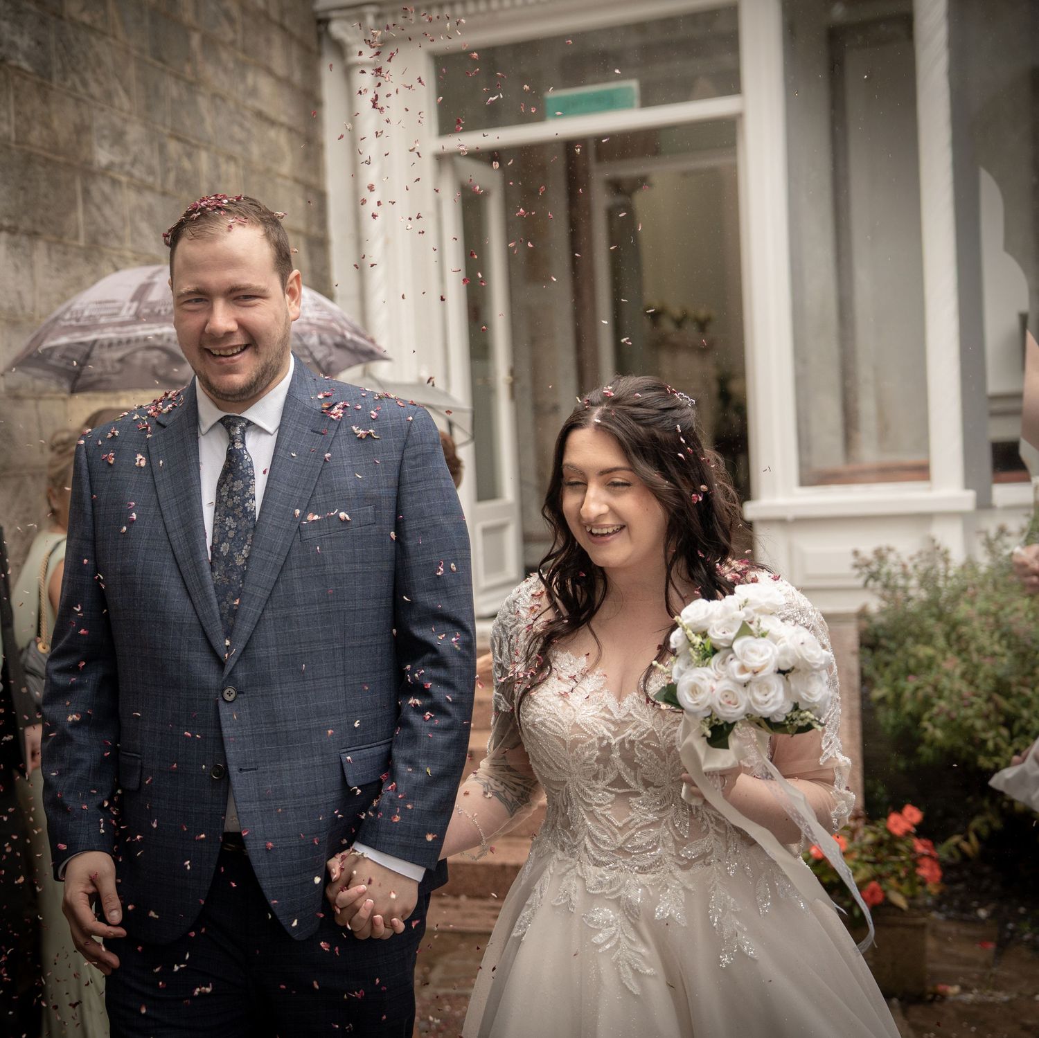 A joyful bride and groom walk hand in hand through a shower of confetti outside a stone building on their wedding day.
