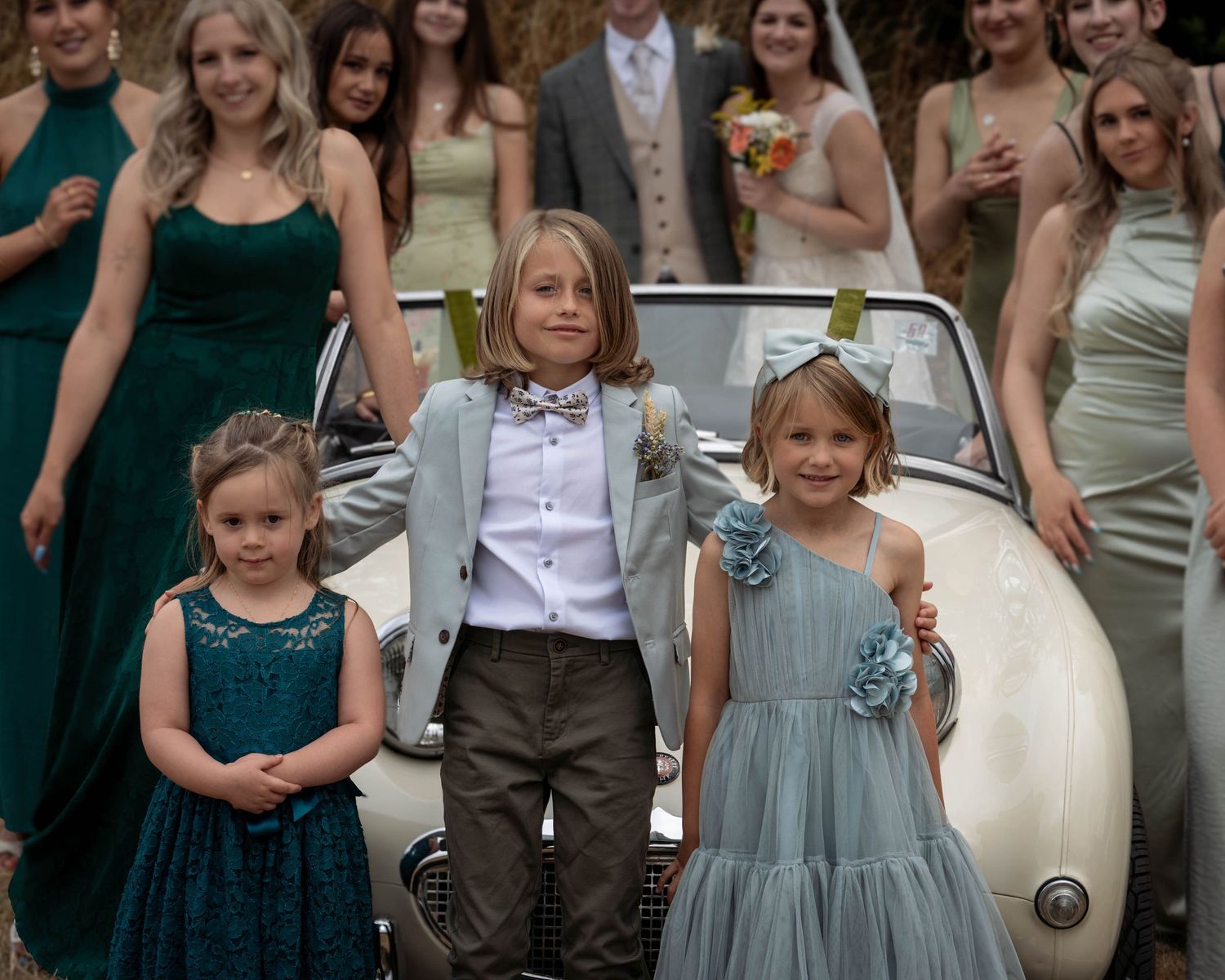 Three children pose in front of a vintage white car surrounded by wedding party members in green and teal formal attire.