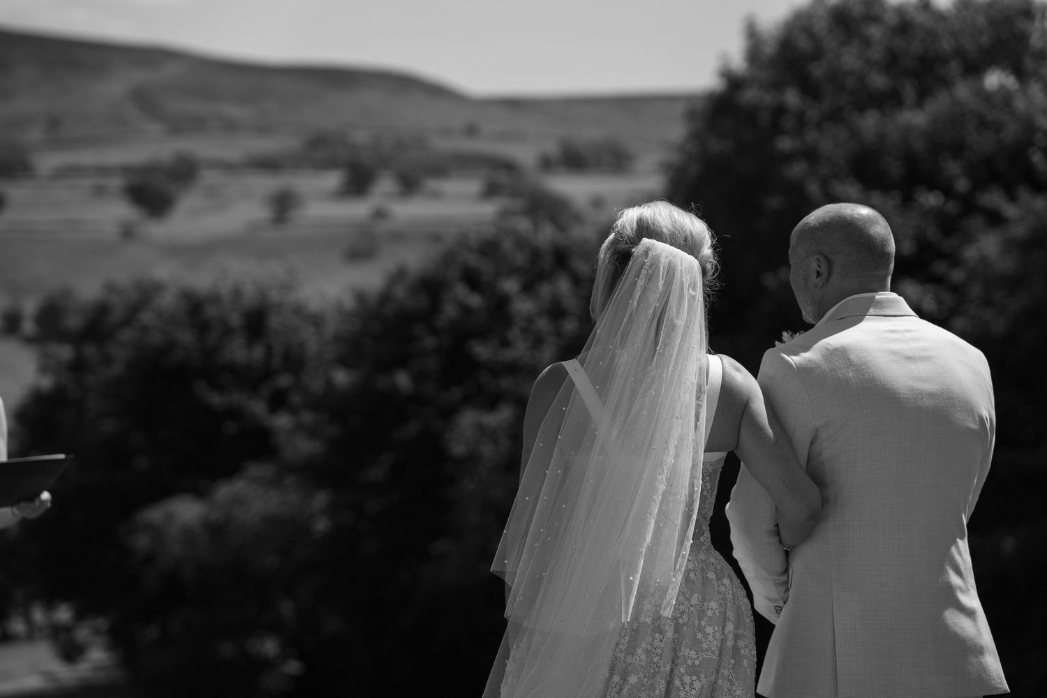 Black and white photo of a bride in veil and groom in suit standing together overlooking a scenic countryside landscape.