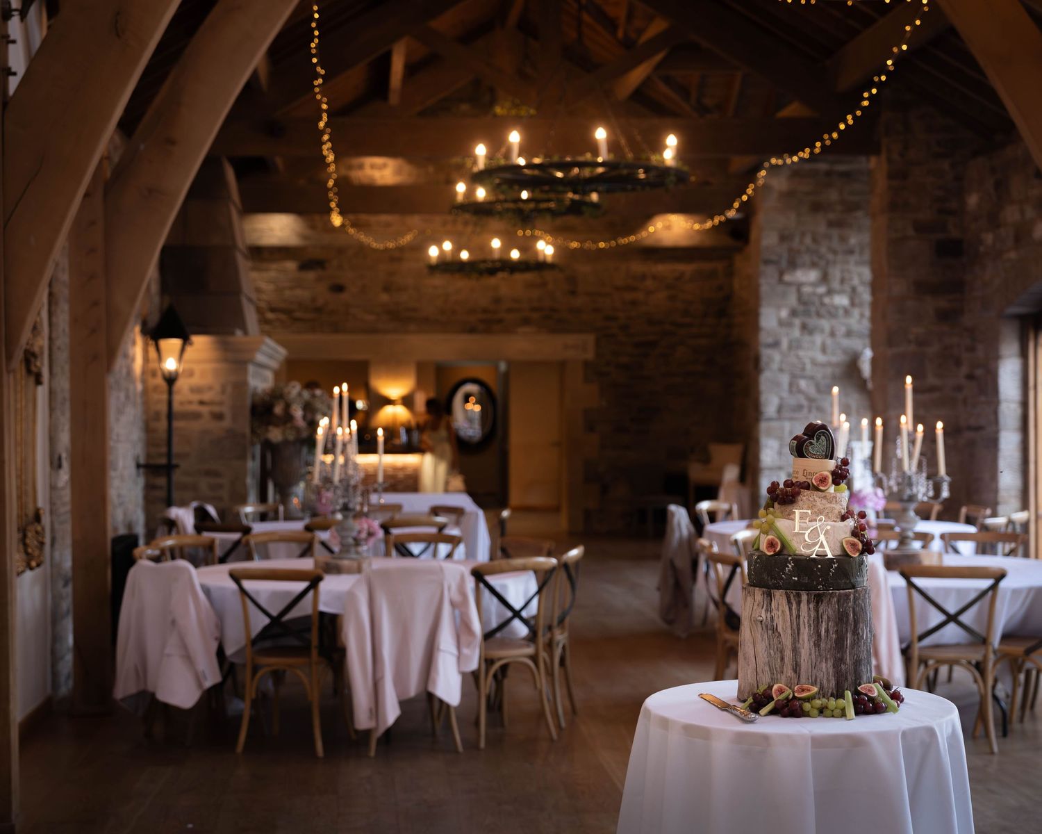 Rustic stone barn wedding reception with candlelit tables, wooden cross-back chairs, and warm fairy lights overhead.
