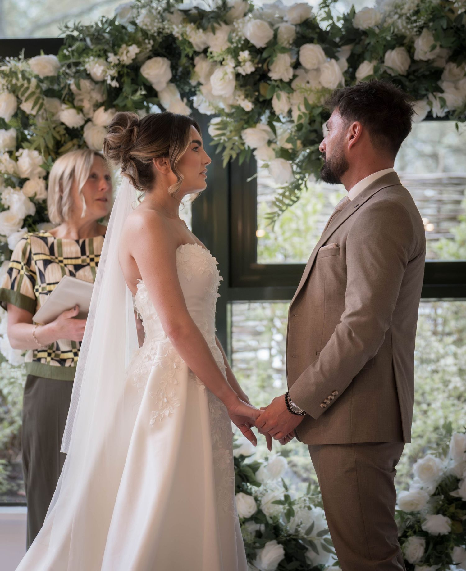 Bride and groom holding hands at floral arch ceremony with white roses, officiant in background.