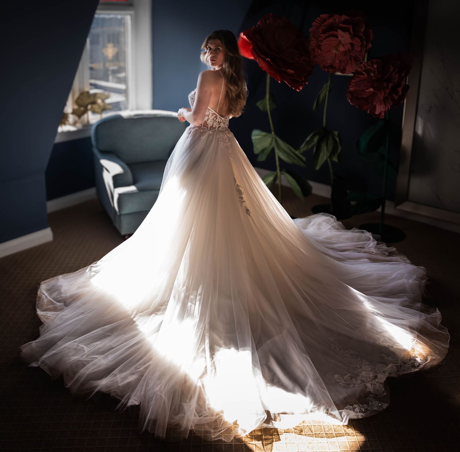 Bride in elegant backless lace wedding gown with flowing tulle train, bathed in natural light near a window.